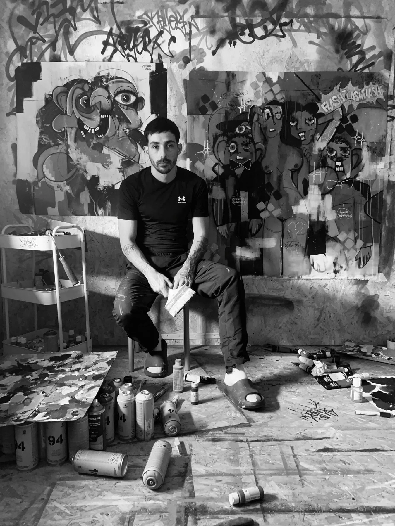 A man sitting in an art studio with paintings and modern artworks on the walls, surrounded by spray paint cans and art tools.