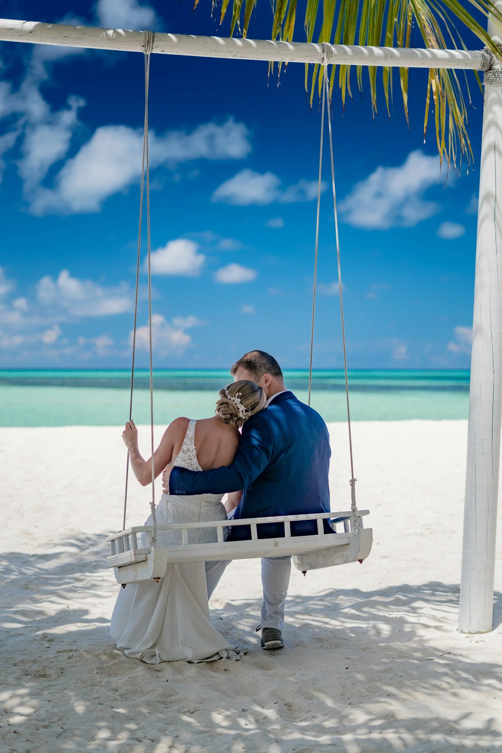A newlywed couple sits on a swing on a sandy beach, sharing an intimate moment under a clear blue sky with some clouds, overlooking the ocean.