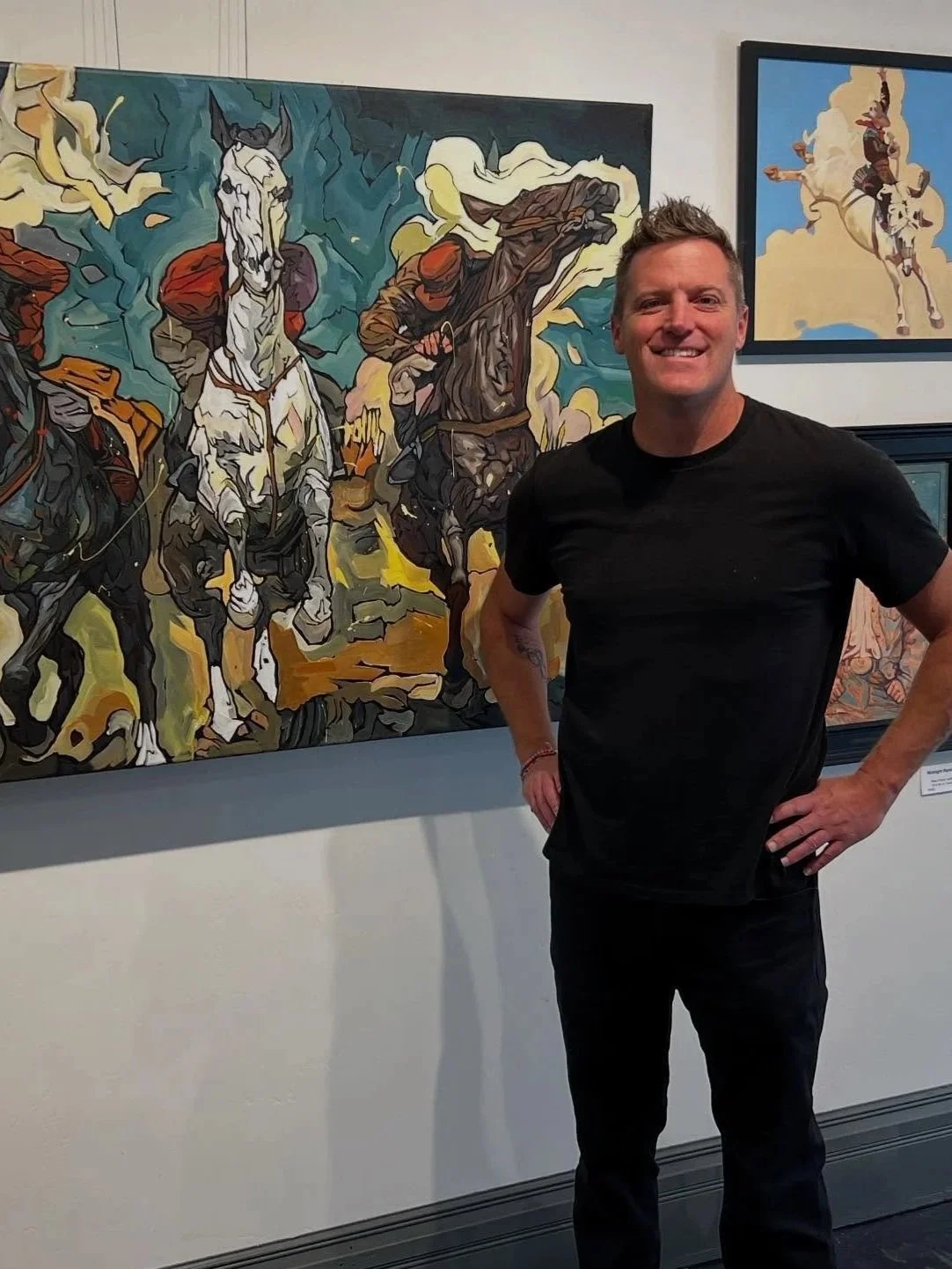 A man smiling in an art gallery, standing next to a large colorful painting of cowboys on horseback.