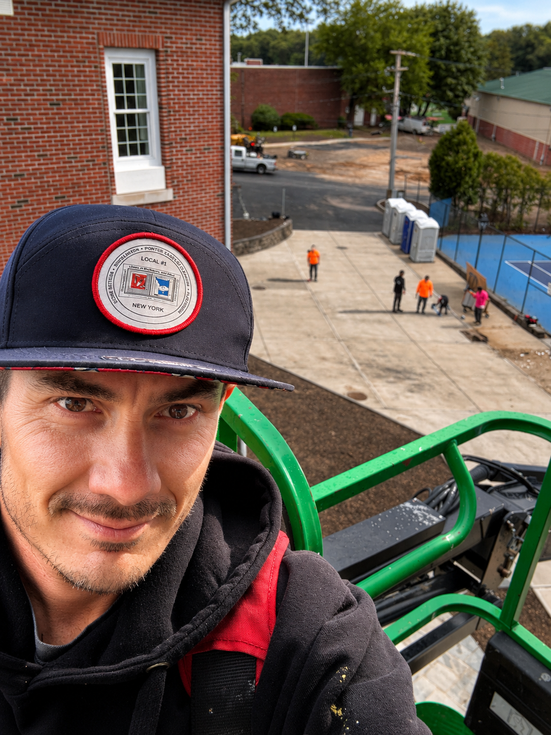 Person in a black cap with a red and white patch, smiling, taking a selfie from a green lift at a construction or maintenance site with workers on the ground, a brick building, a tennis court, trees, and portable toilets in the background.