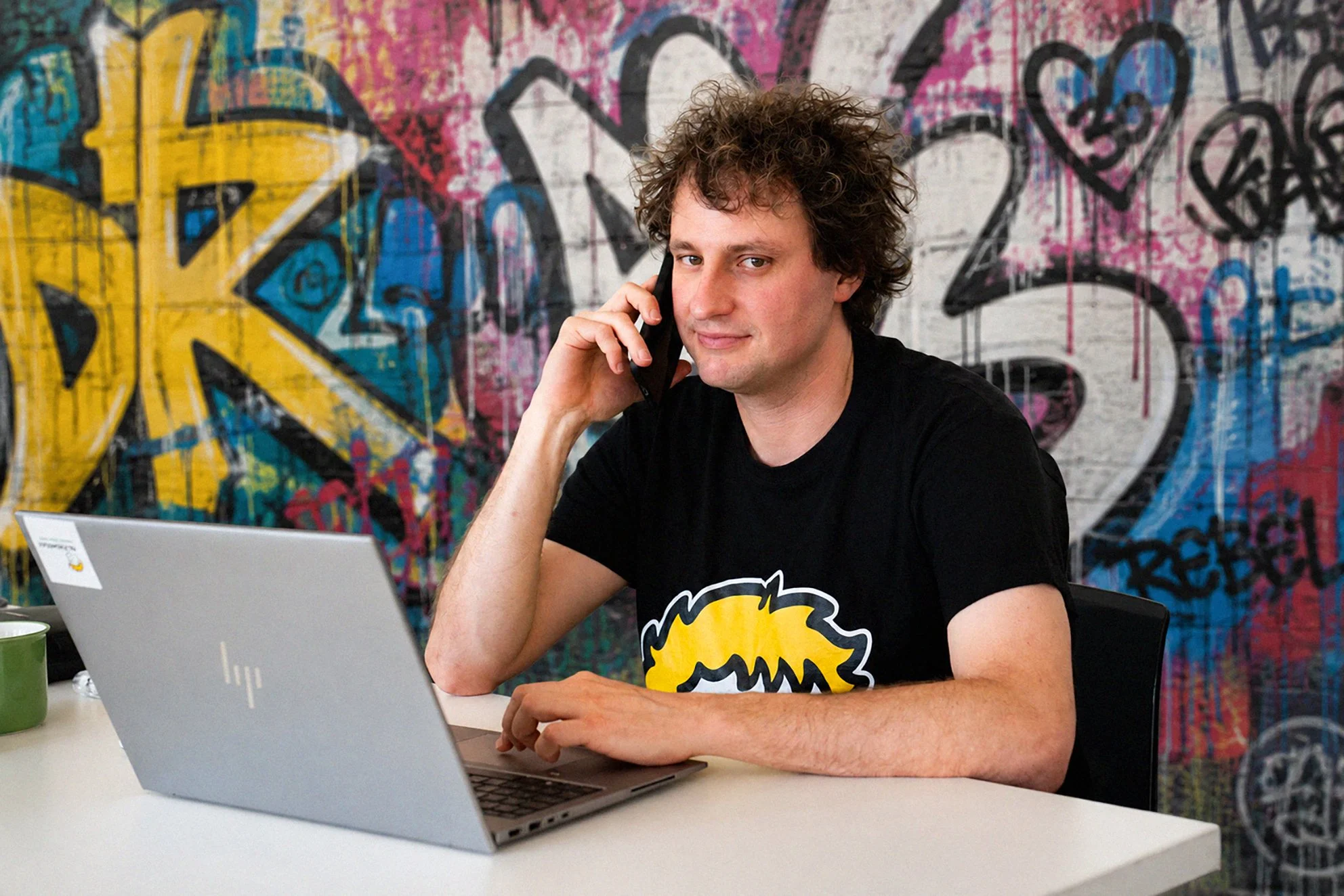 Michael Schmelcher from Re:Rebellion sits in front of his graffiti wall and looks slyly into the camera.