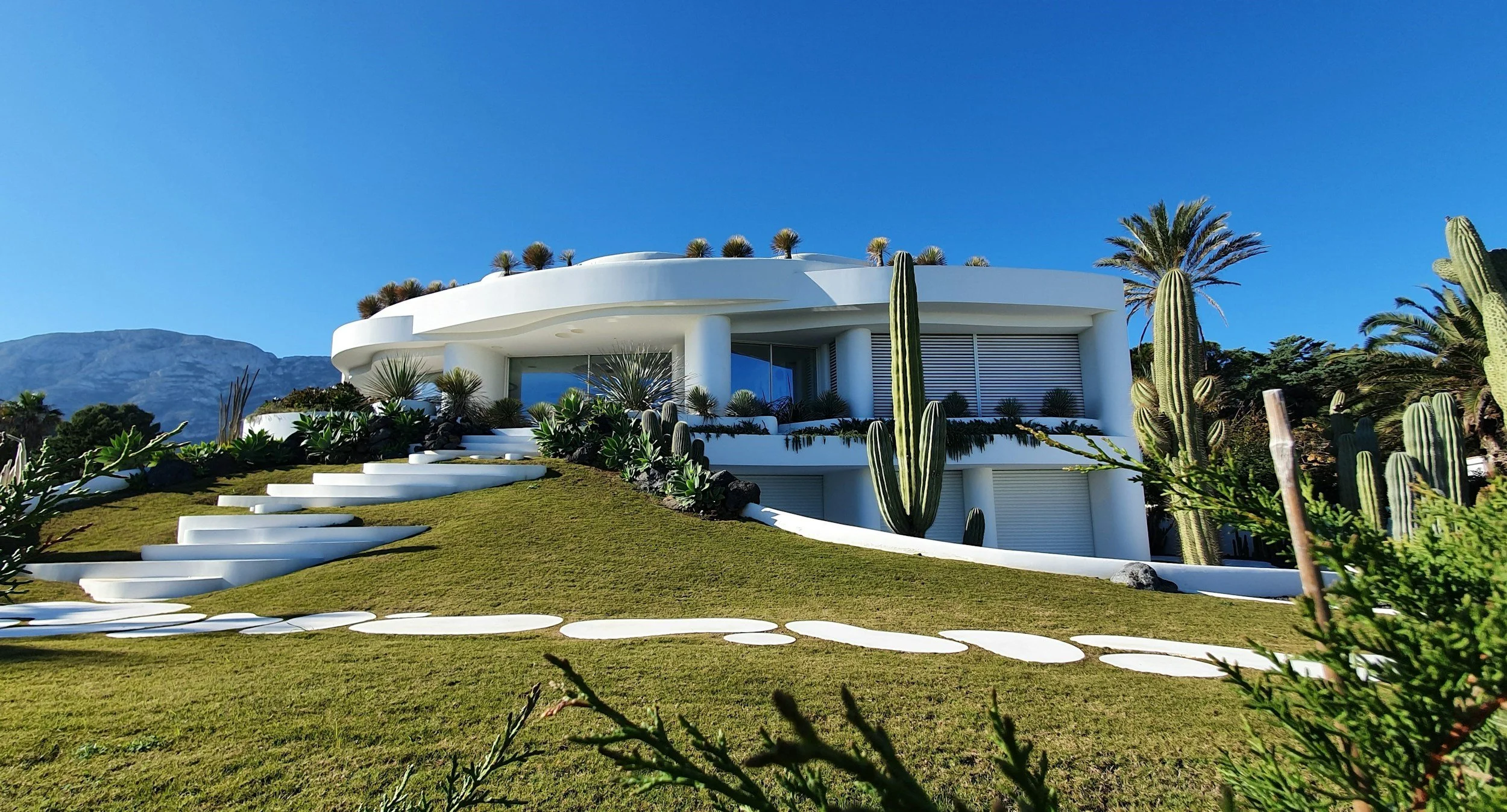 Modern white house with curved architecture, surrounded by desert plants and cacti, on a grassy hill against a clear blue sky.