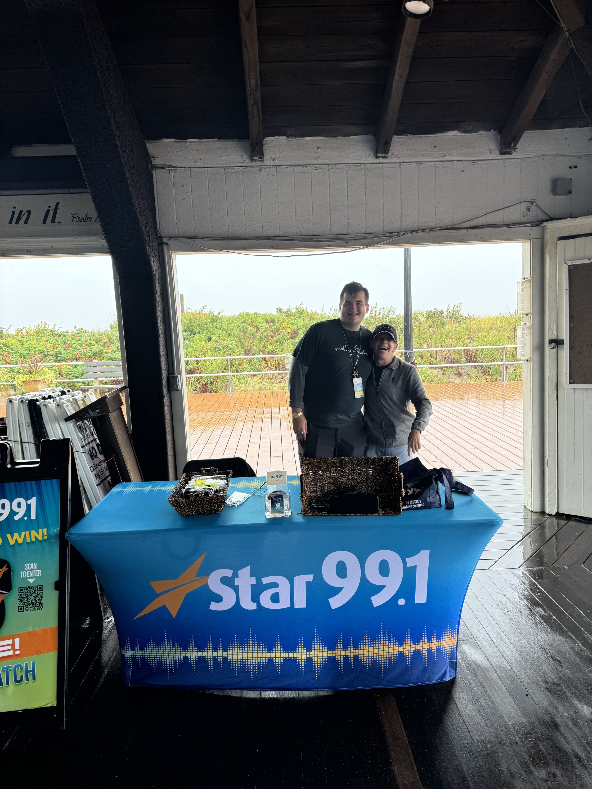 Two smiling people standing behind a Star 99.1 radio station table inside a building with large open windows showing a outdoor terrace and greenery.