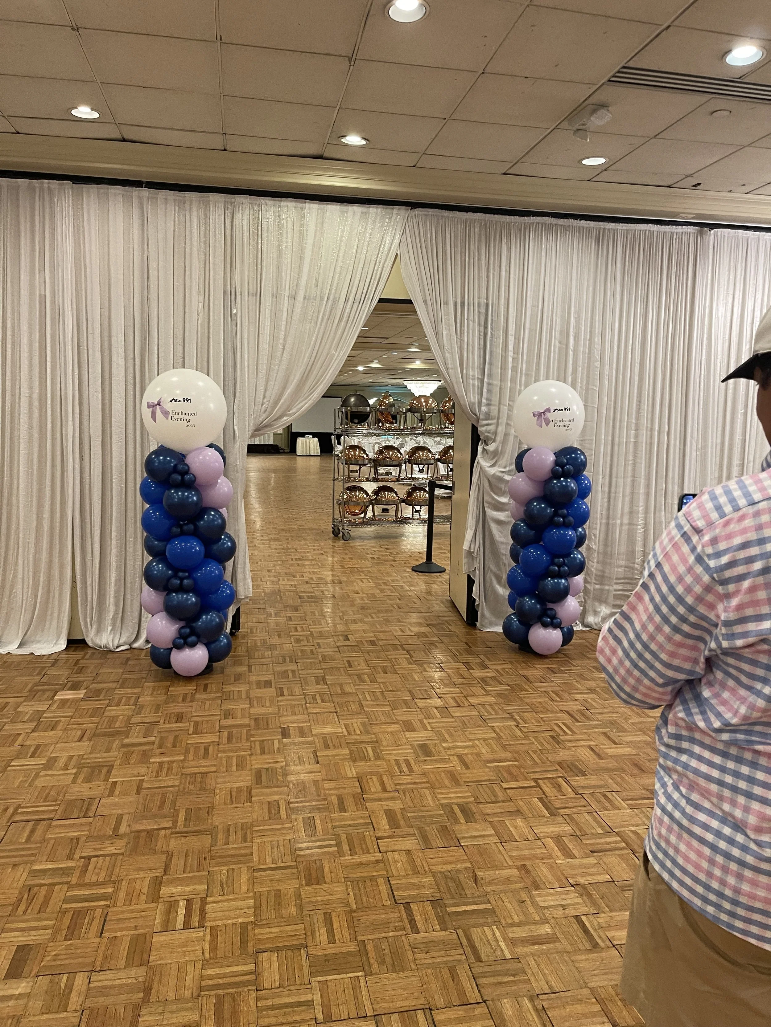 Decorated entrance to event space with white curtains and balloon columns with blue, purple, and dark blue balloons.