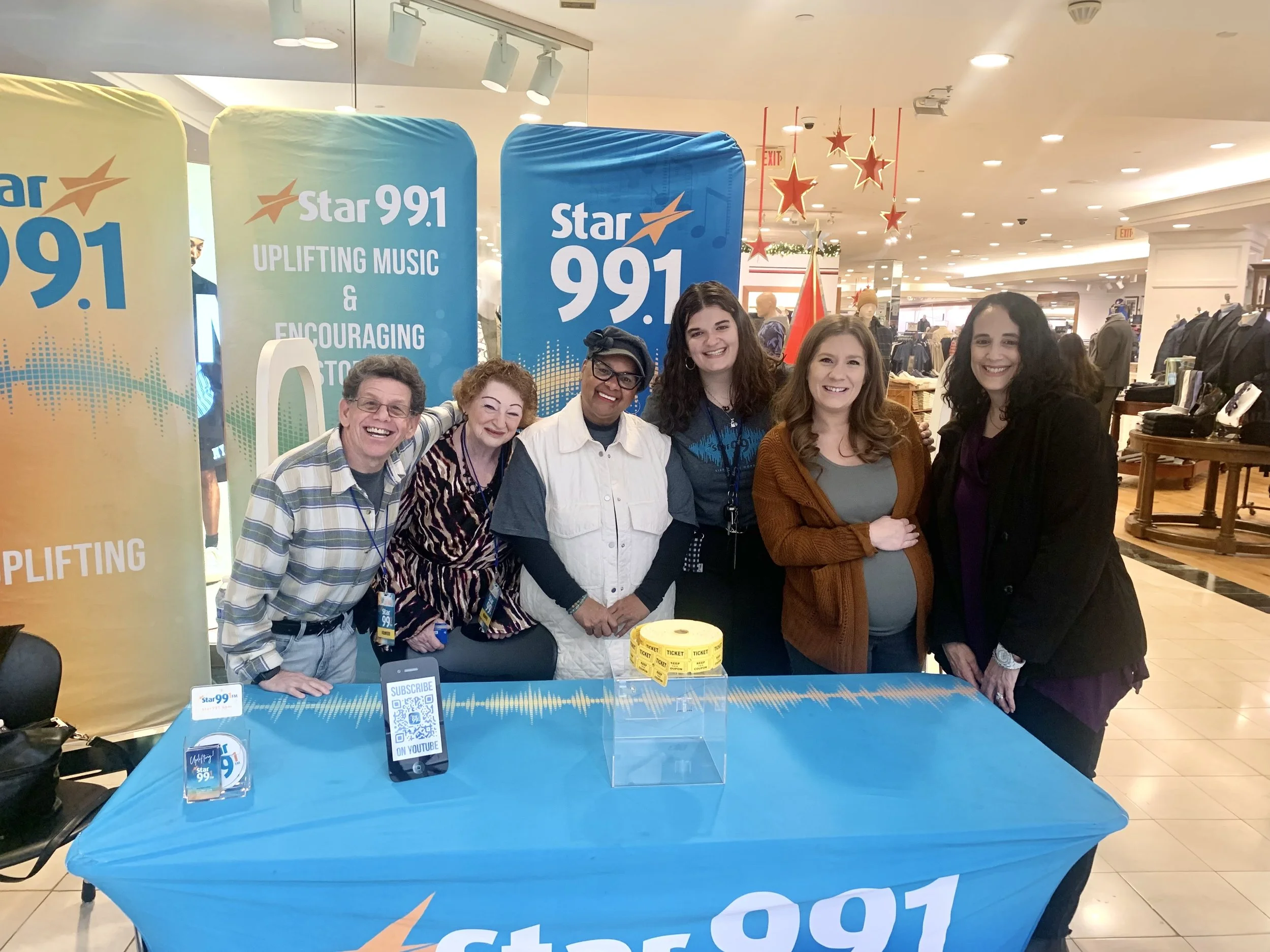 Group of six people standing behind a table with a Star 99.1 banner in a shopping mall, smiling for a photo, with Christmas decorations hanging overhead.