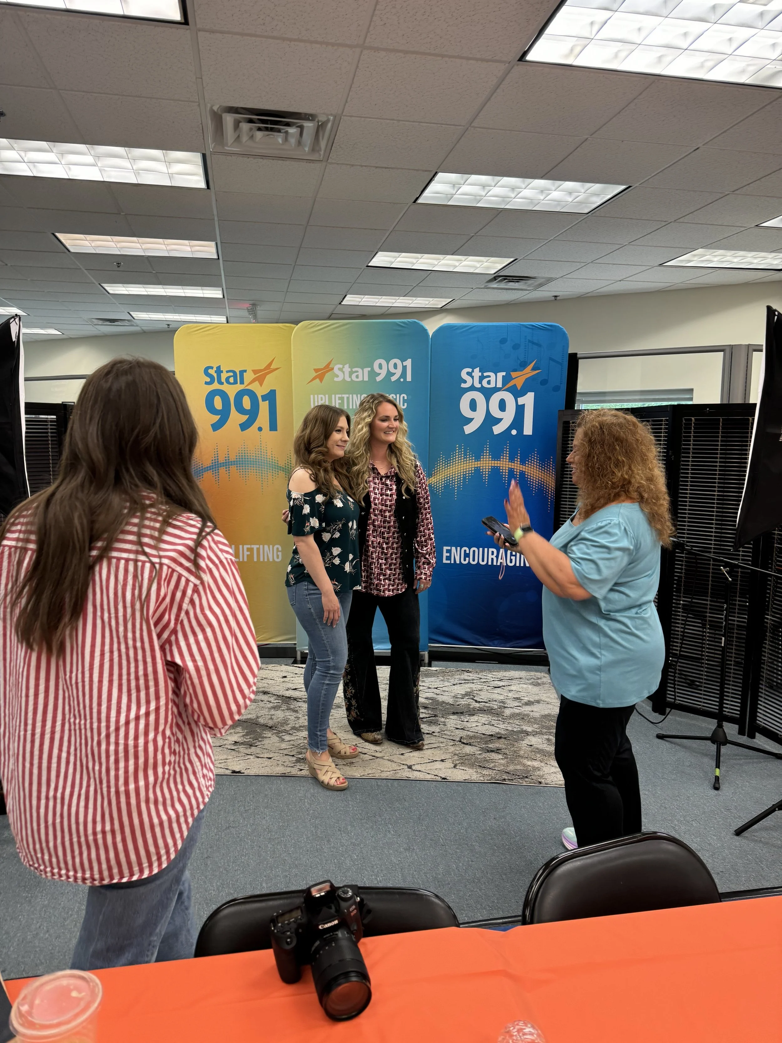 Two women standing in front of a Star 991 backdrop, being filmed or photographed by a woman with curly red hair, in an office or conference room setting.