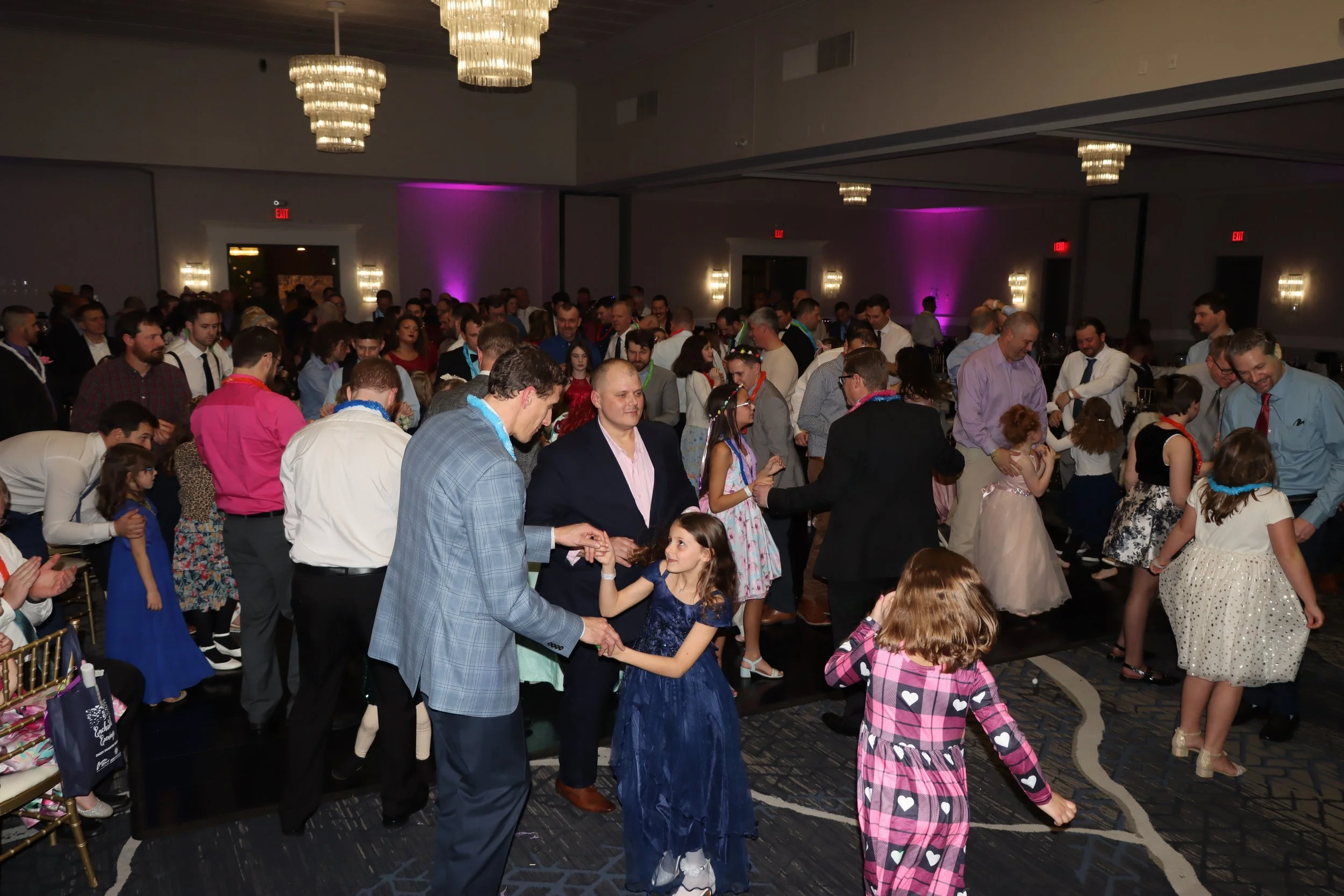 People dancing at a celebration or party in a large indoor venue with elegant chandeliers and purple lighting.