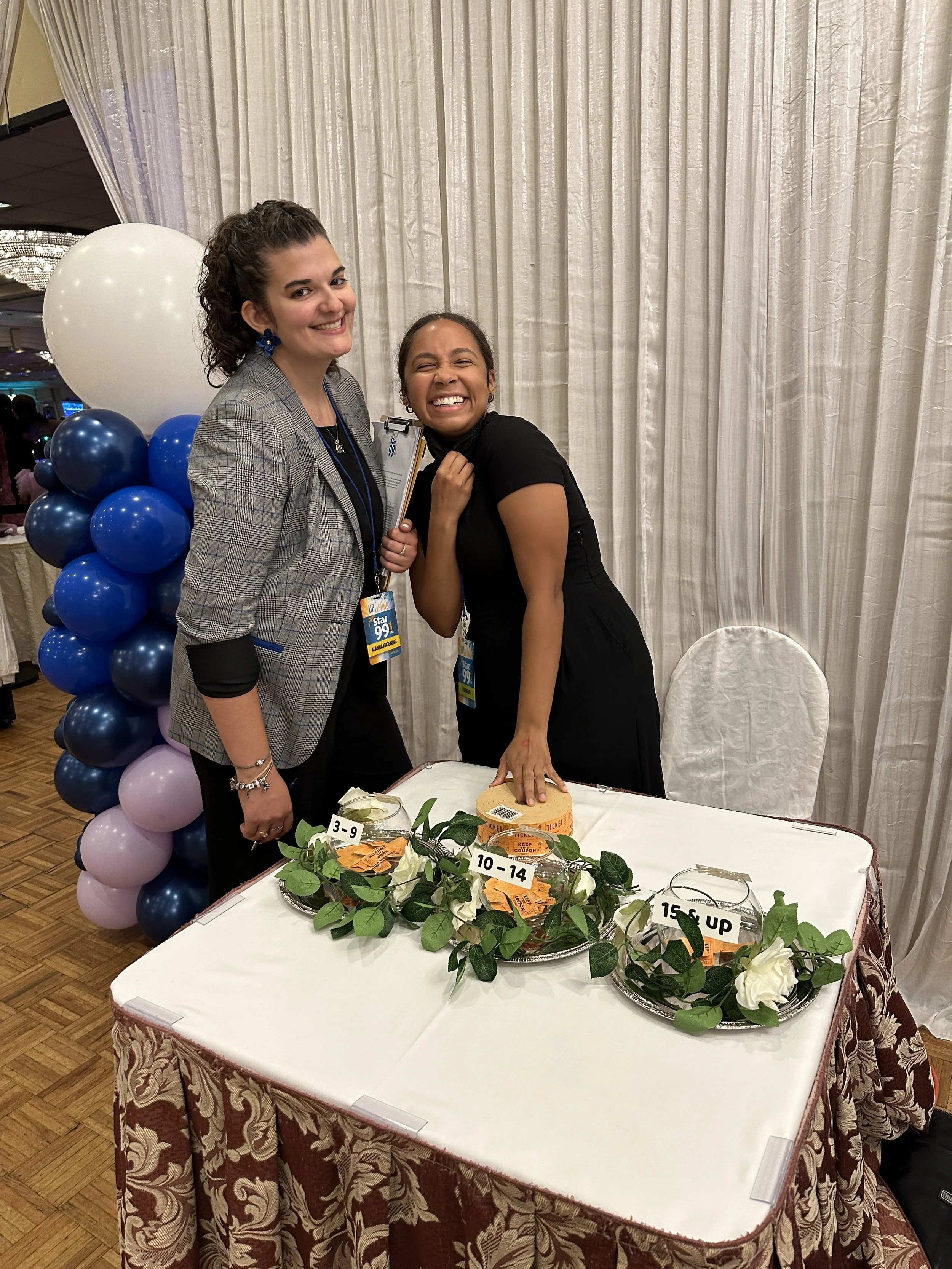 Two women smiling and posing behind a table with raffle tickets and flowers at an indoor event. One woman has curly dark hair and is wearing a blazer, the other has straight dark hair and is wearing a black dress. Behind them is a white curtain and b