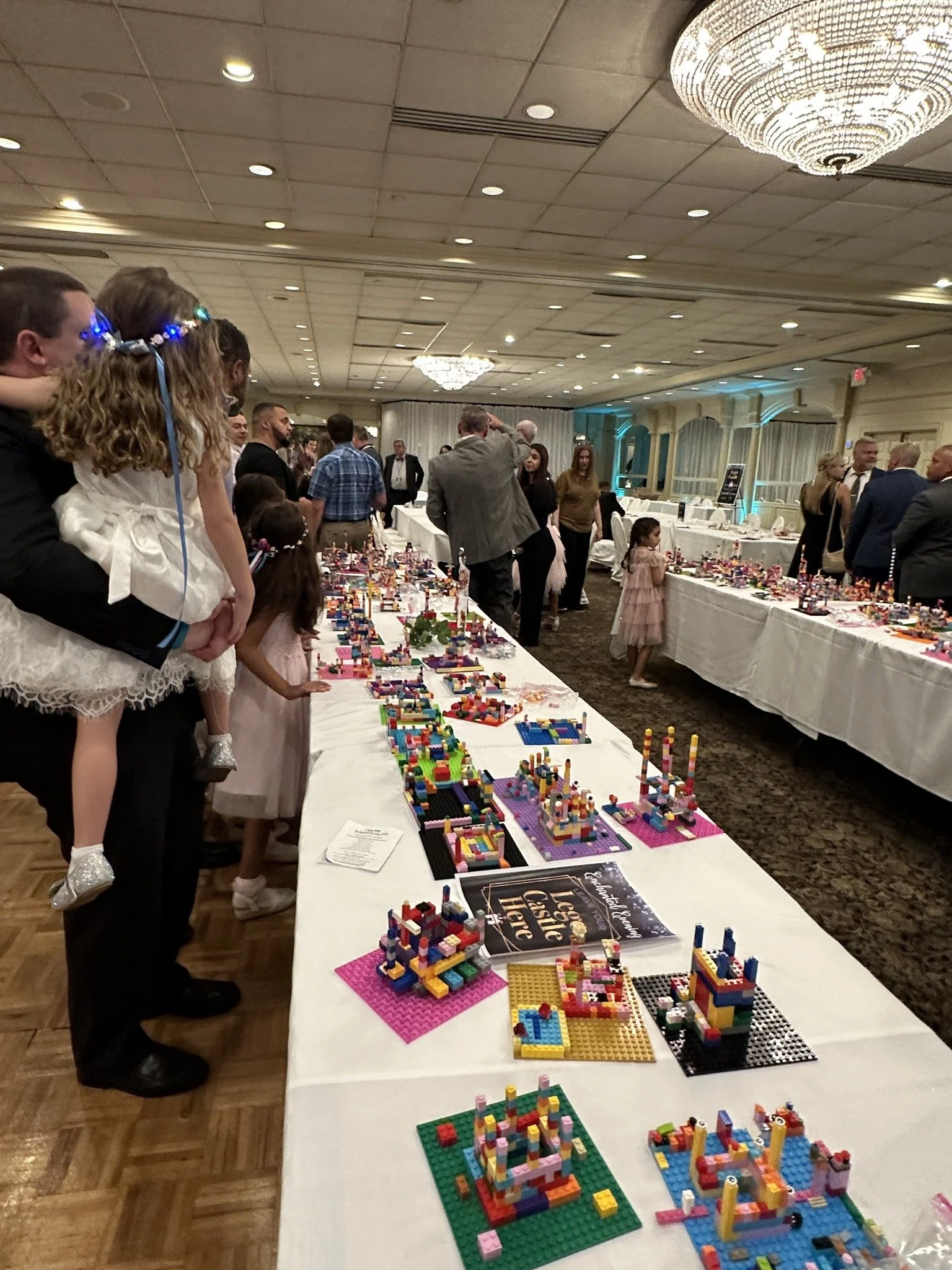 People at a collection of Lego displays at an event with a white tablecloth, including children and adults, in an elegant hall with chandeliers and decorative lighting.