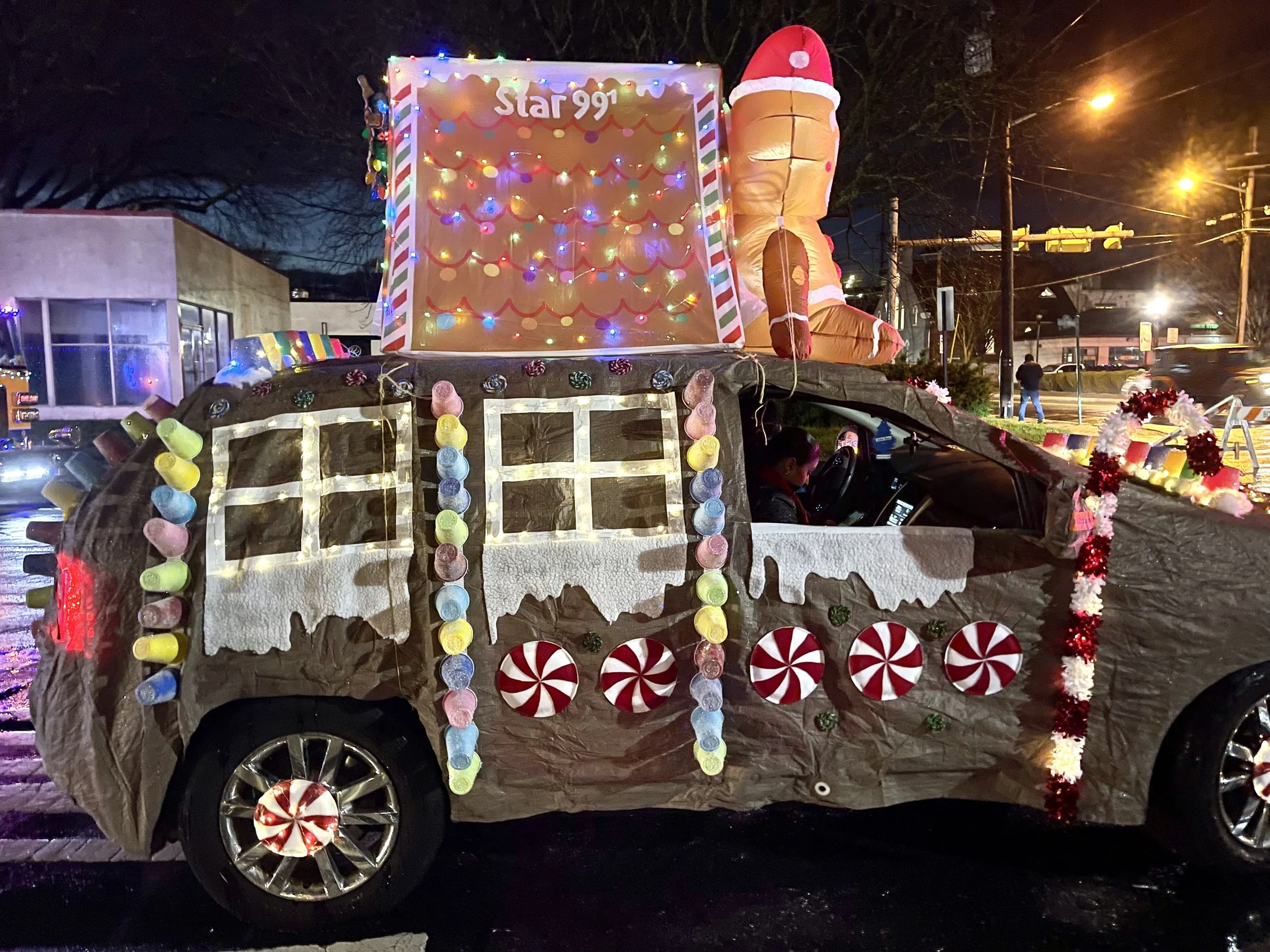 A car decorated as a gingerbread house with Christmas lights, peppermint candy decorations, and an inflatable gingerbread man with a Santa hat on top.
