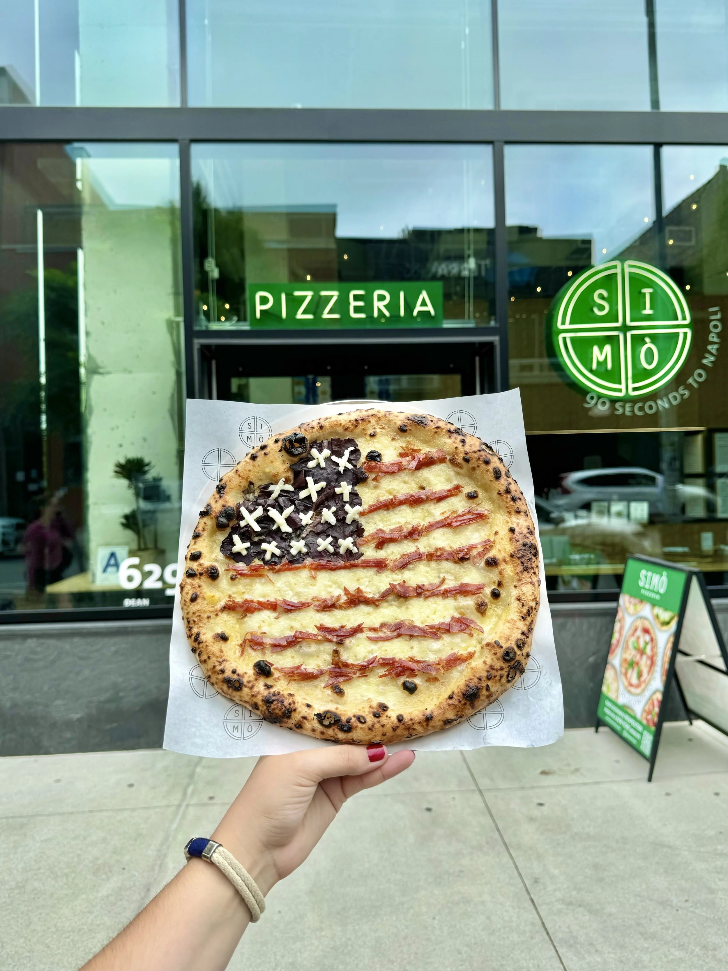 A pizza with cheese, black olives, and sliced cured meat toppings styled like the American flag, presented outside a pizzeria called SIMÒ Pizza. 