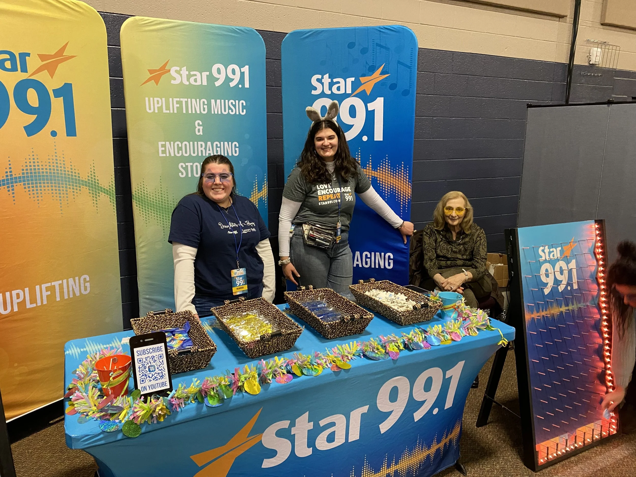 Three women at a Star 99.1 radio station promotional event booth, with one woman wearing bunny ears, standing behind a table with Easter-themed decorations and trays of baked goods, against a backdrop with star 99.1 branding.