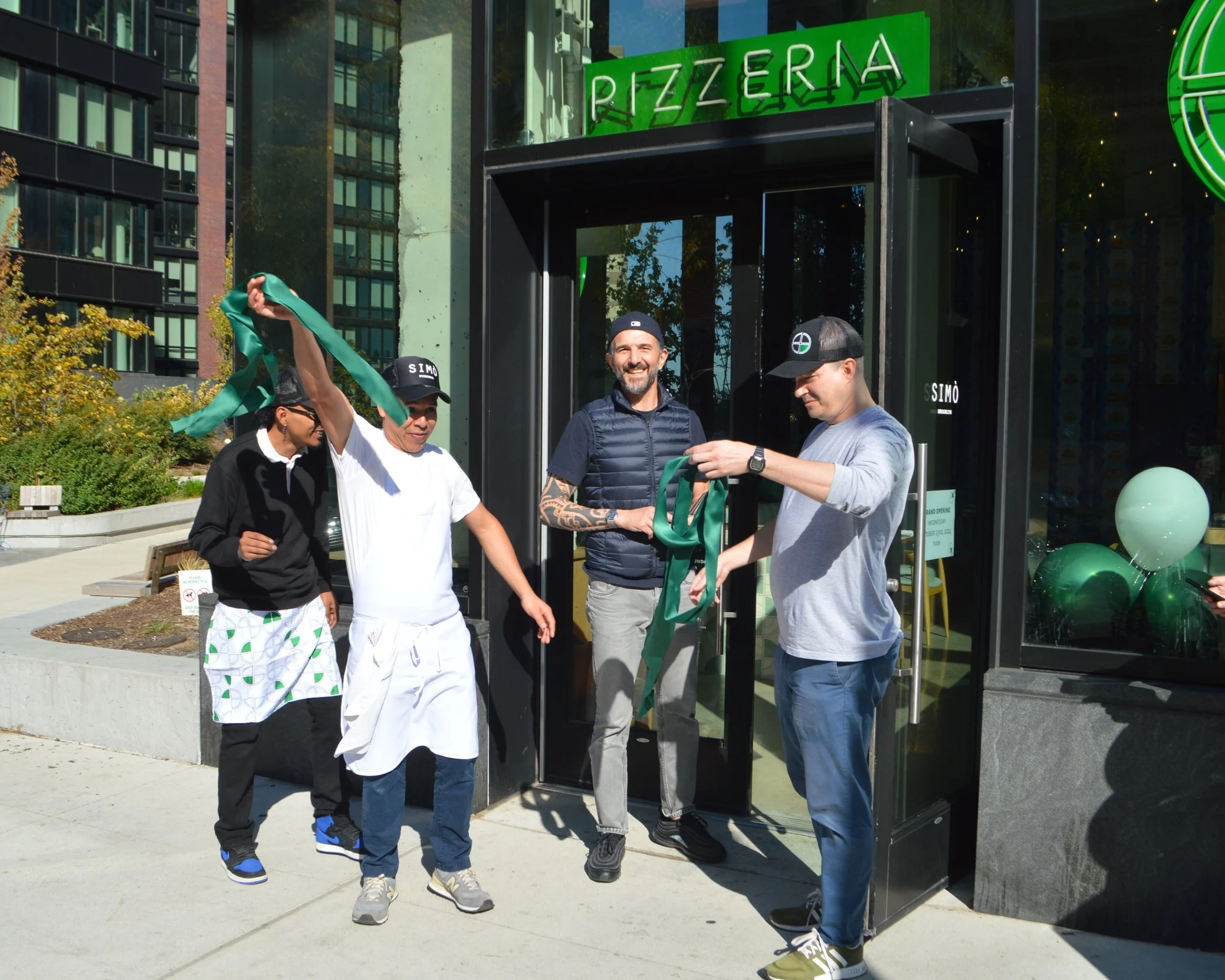 Group of four people celebrating outside a restaurant with a green neon sign that says 'PIZZERIA'. They are holding and exchanging green ribbons, and one person is wearing an apron. The scene suggests a ribbon-cutting or opening event.
