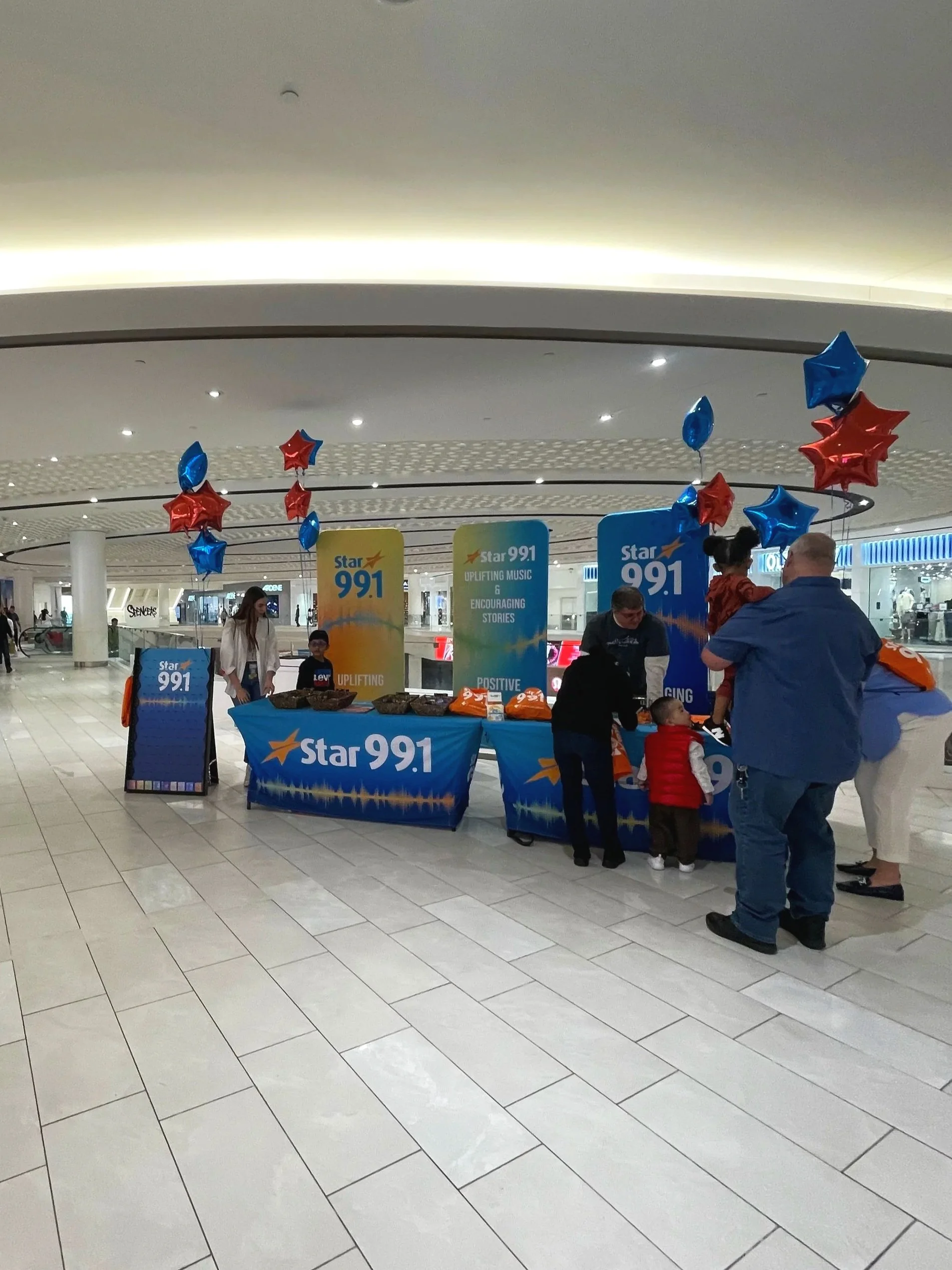 A promotional booth for Star 99.1 radio station at an indoor mall, decorated with red and blue star-shaped balloons. Several people, including children, are interacting at the booth which has banners and tables with promotional materials.