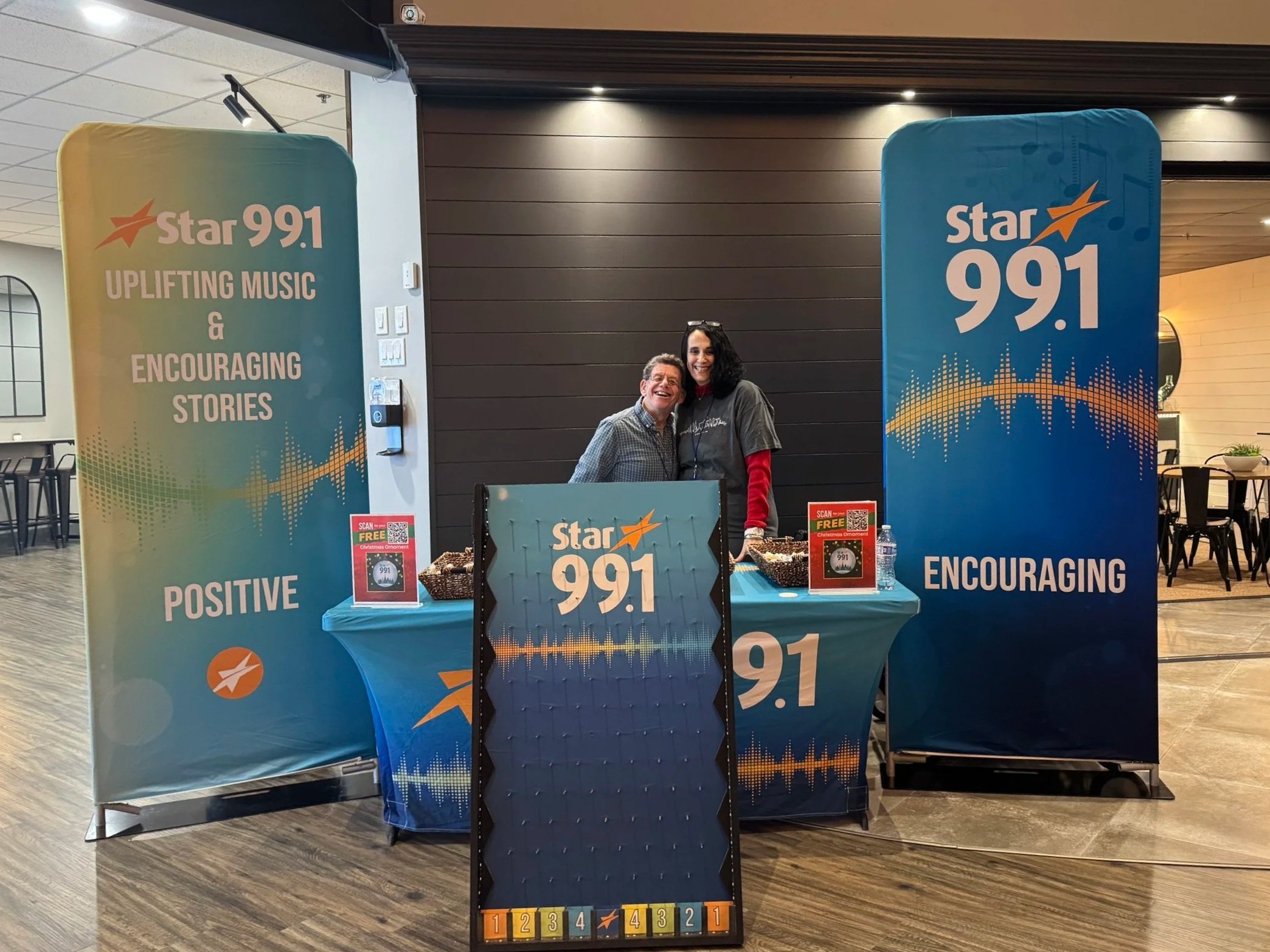Two people standing behind a table at a promotional booth for Star 99.1 radio station, with banners displaying the station's logo, frequency, and positive message, in an indoor setting.