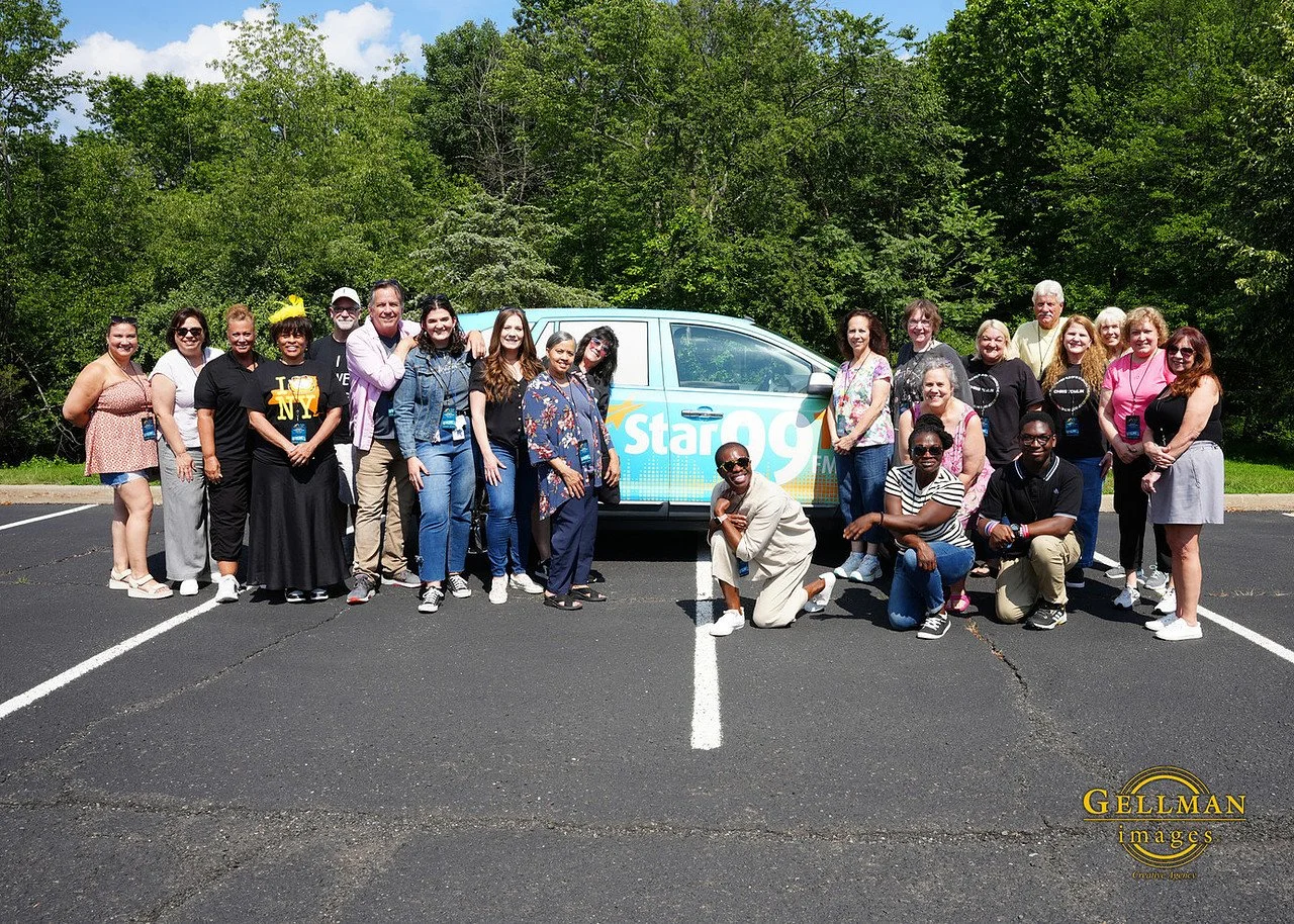 A group of 21 diverse people posing for a photo in a parking lot with a Star 99 FM vehicle behind them, surrounded by trees.