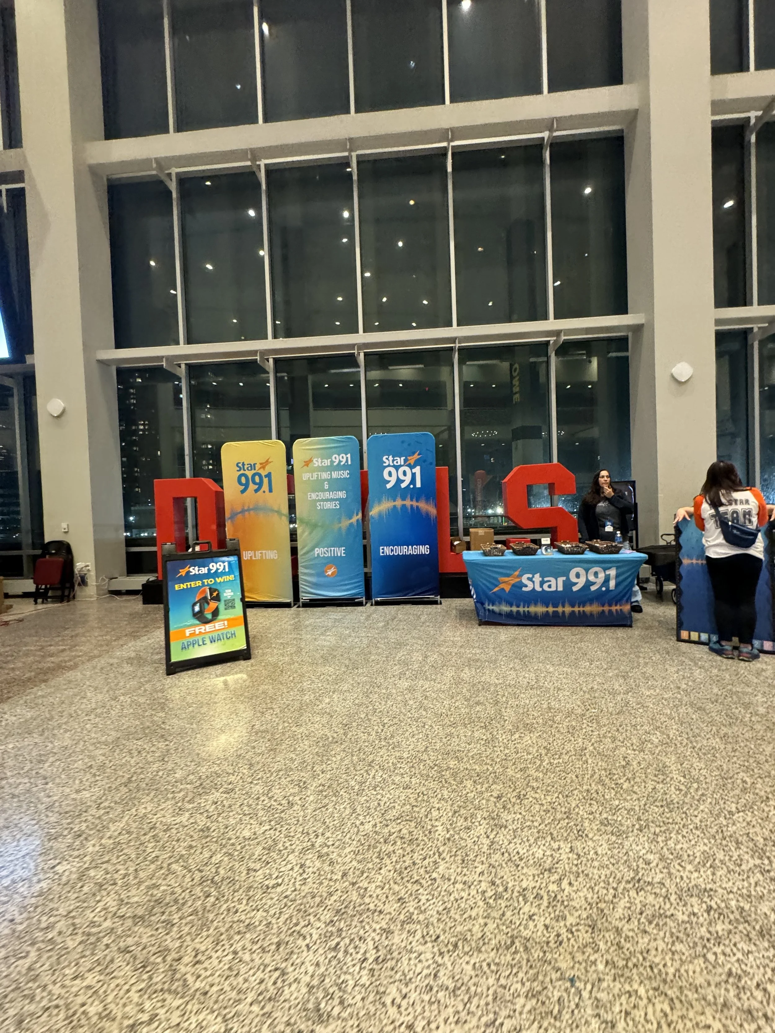 An indoor promotional event for Star 99.1 radio station featuring banners, a table with promotional items, and a sign advertising a contest to win a free Apple Watch.