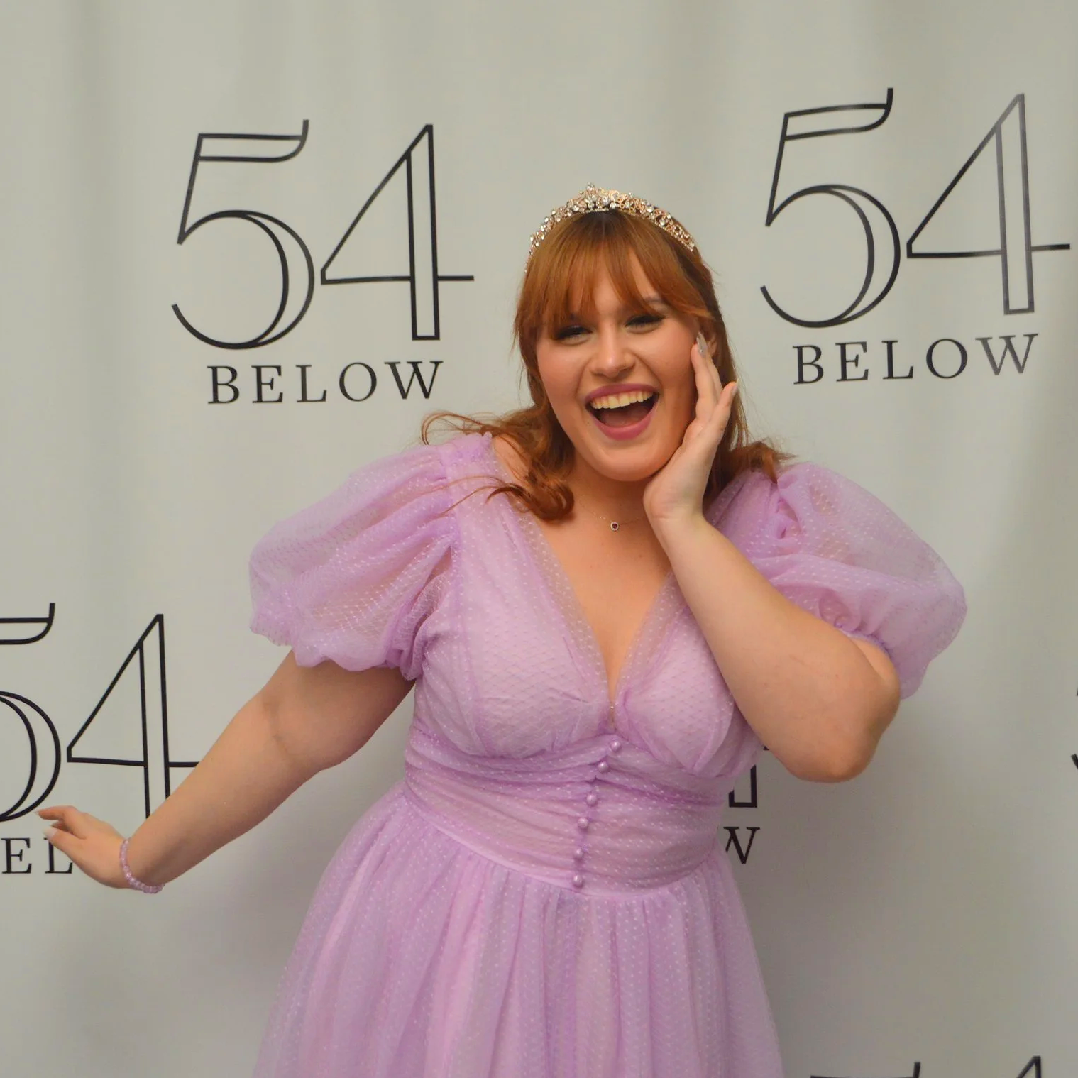 A woman in a pink dress and tiara smiling and posing in front of a backdrop with the numbers 54 and the word BELOW.