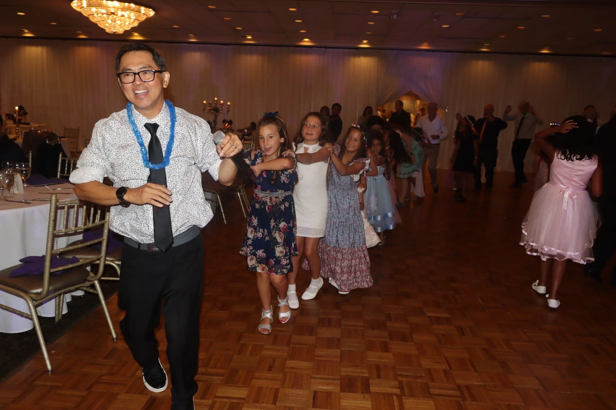 A man leading a conga line of children at a party or celebration in a large decorated hall.