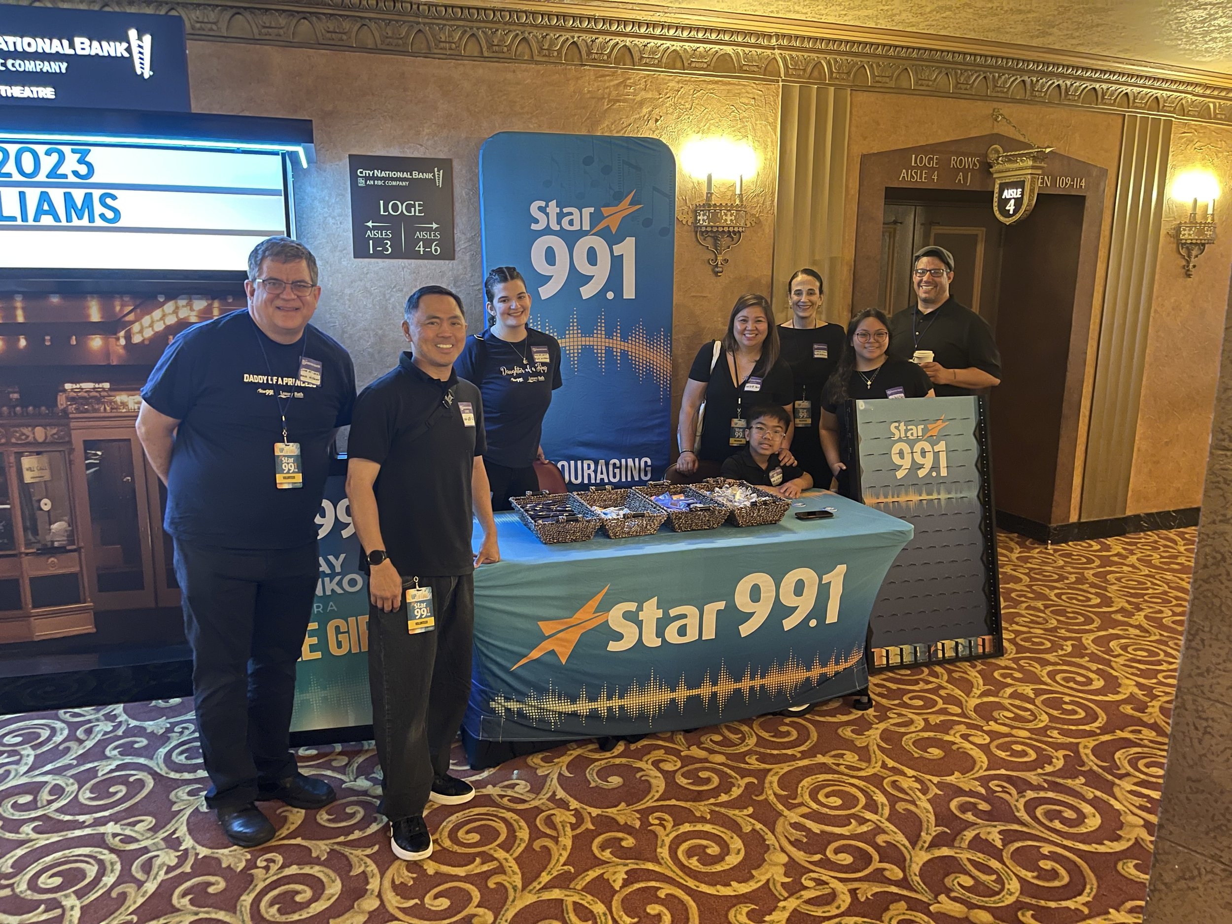 A group of seven people standing behind a table with Star 99.1 branding, smiling at the camera at a radio station event in a theater lobby with ornate decor and patterned carpet.