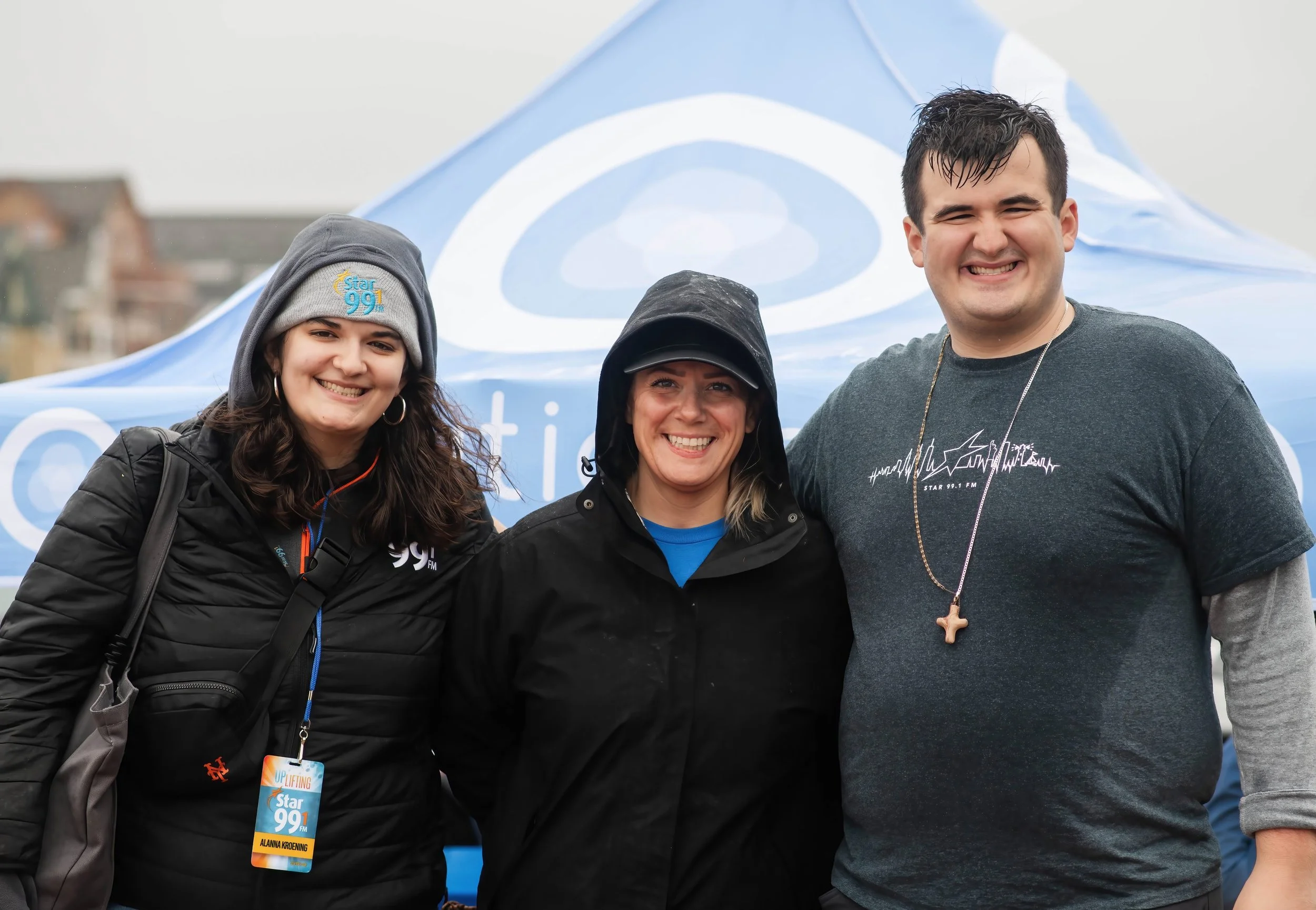 Three smiling people standing outdoors in front of a blue tent, wearing casual clothing and jackets. The person on the left is a young woman with dark hair, wearing a gray beanie and a black jacket with a "Star 99.1" badge. The middle person is a wom
