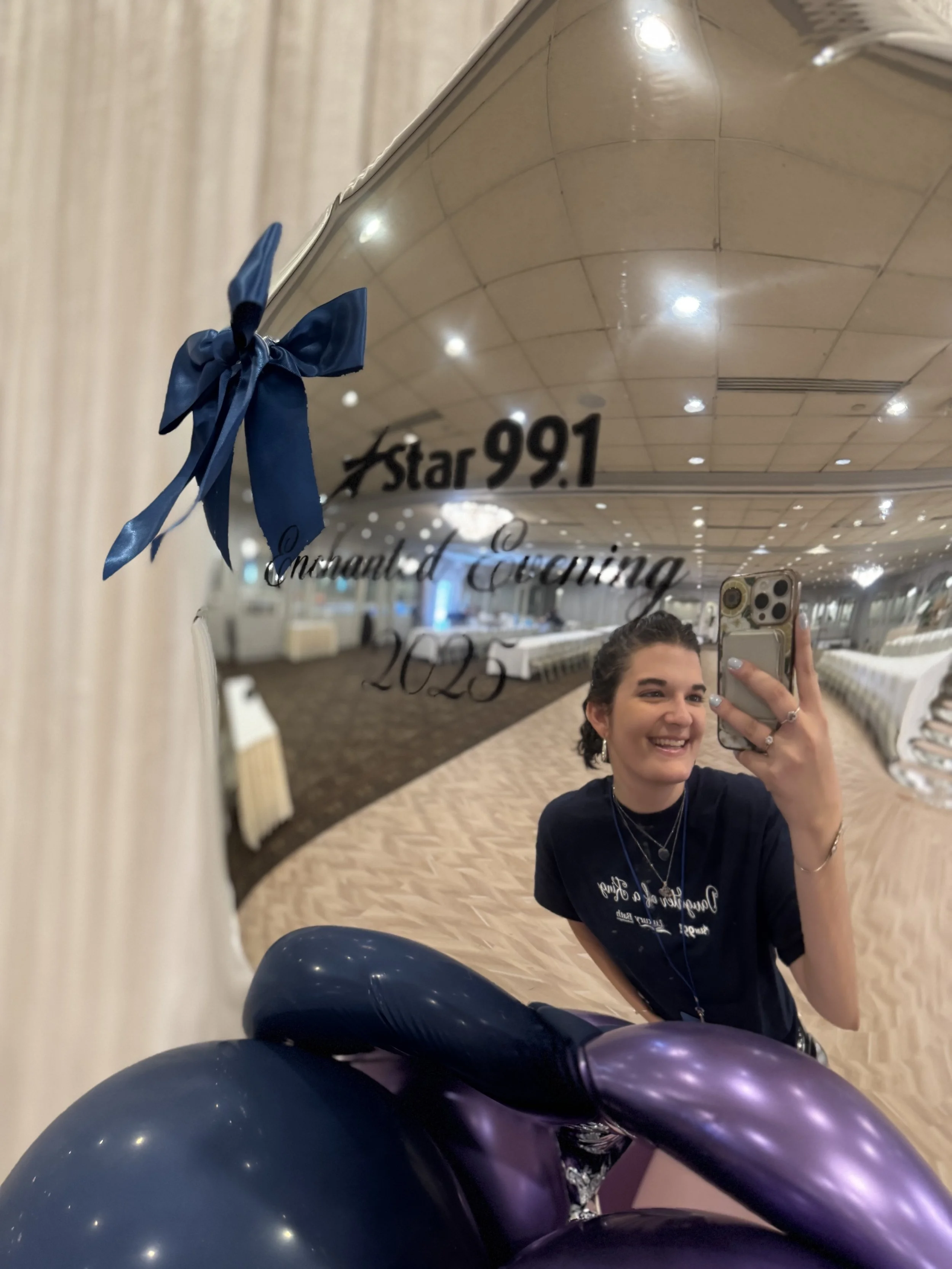 A woman taking a selfie in a reflective balloon sculpture with the inscription 'Star 99.1, Daddy/Daughter Dance 2025,' smiling, wearing a dark T-shirt and jewelry, in a decorated indoor space.
