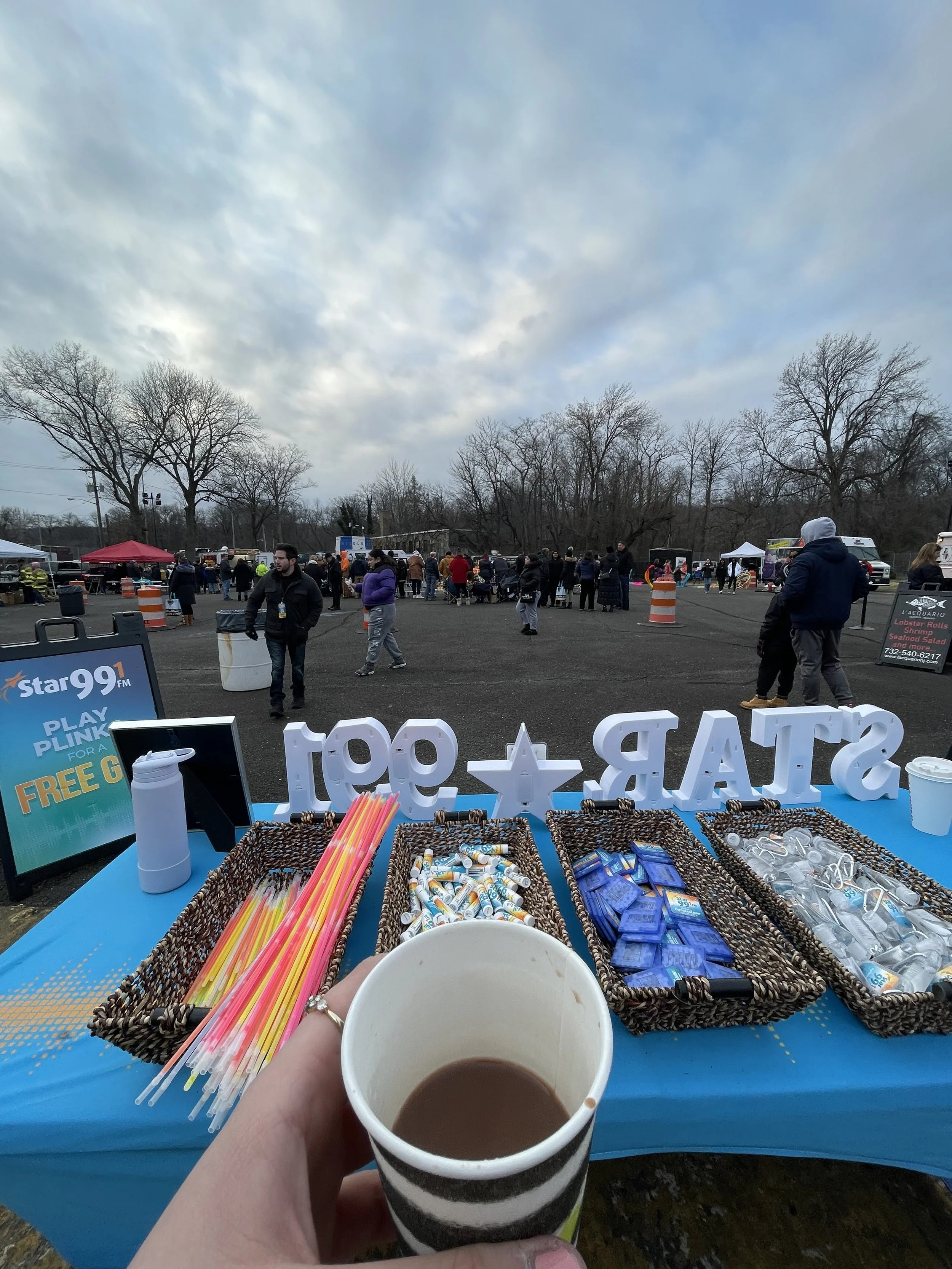 People attending an outdoor event with booths and tents under a cloudy sky. A table in the foreground holds straws, lip balm, and R-Star + 911 signs, with a hand holding a cup of hot chocolate.