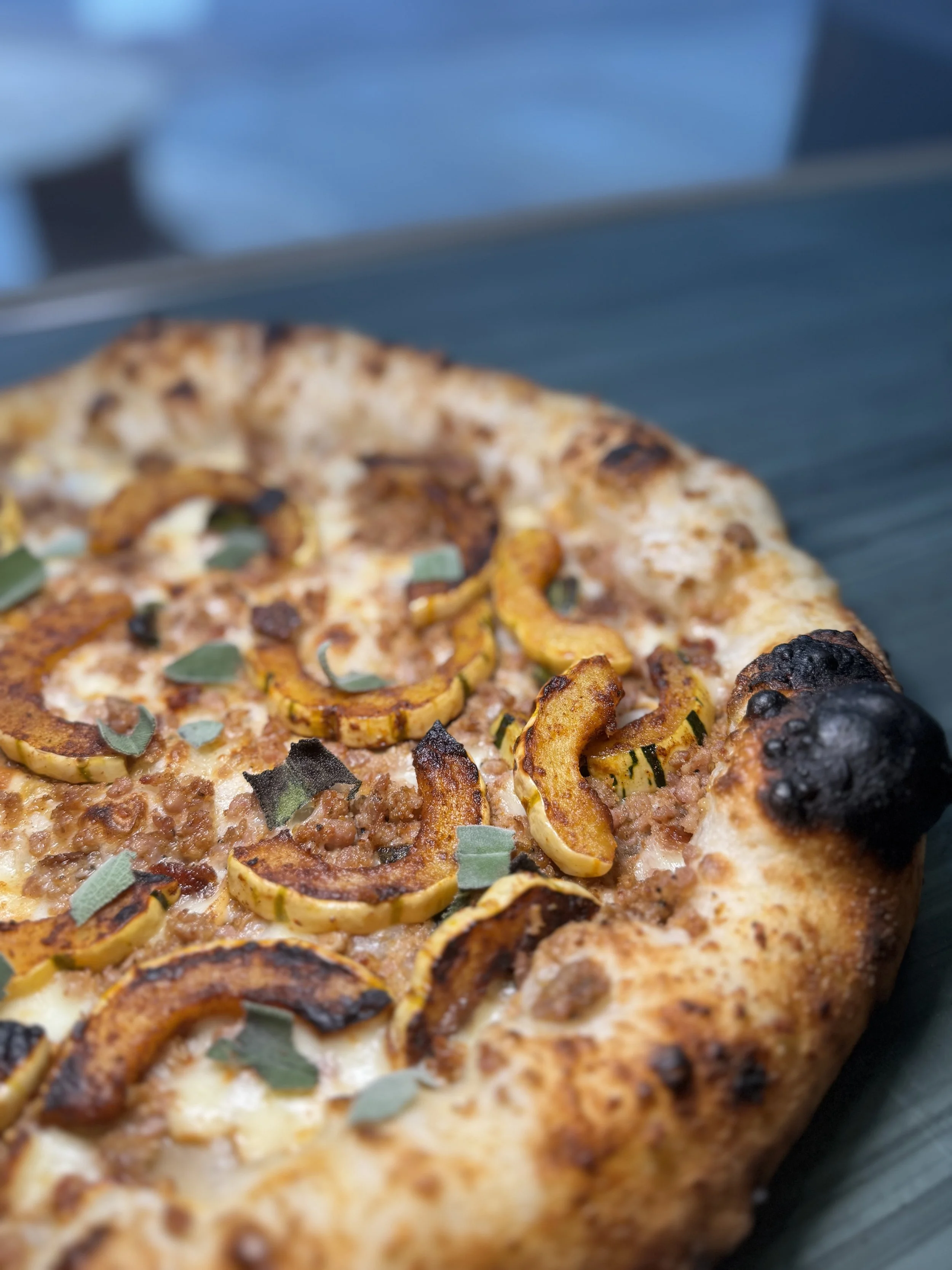 Close-up of a pepperoni pizza with cheese, sliced yellow squash, and sage leaves on a dark green tray.