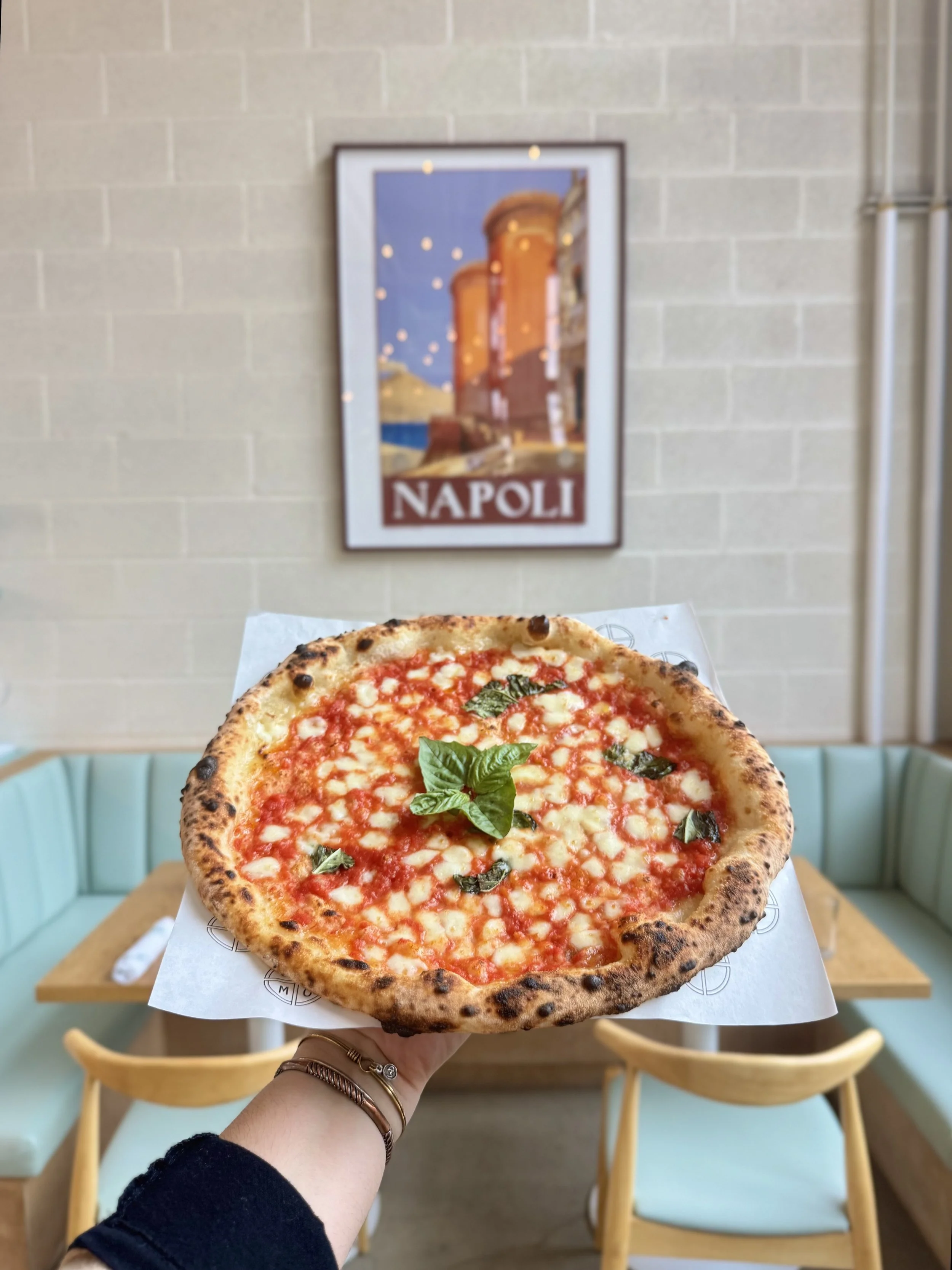 Person holding a Margherita pizza with basil leaves in a restaurant, with a Napoli poster on the wall in the background.