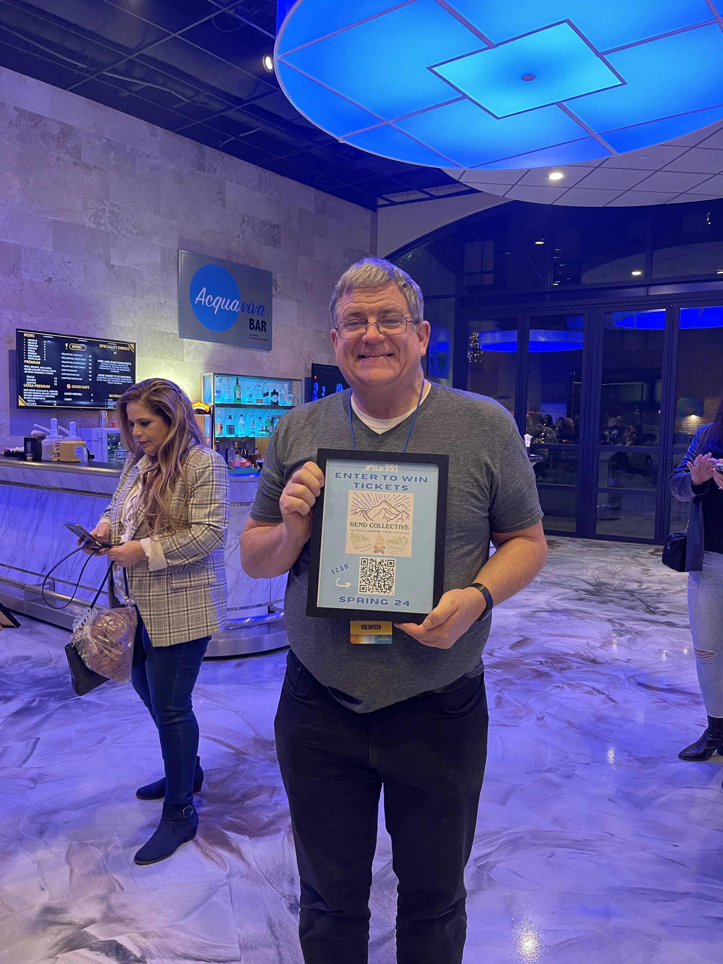A smiling man in glasses holding a framed sign that says 'Enter to Win Tickets' with a QR code and April 24 date at an indoor event, with two women and a bar in the background.