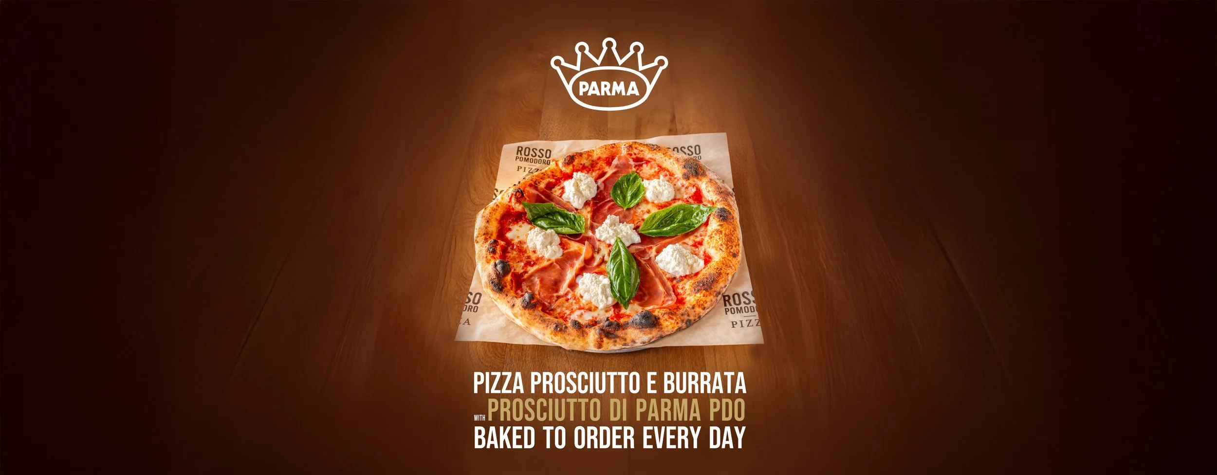 A prosciutto and burrata pizza on a wooden table with basil leaves, placed on a parchment paper with Parma branding. The text overlay states: "Pizza prosciutto e burrata with prosciutto di Parma PDO baked to order every day."