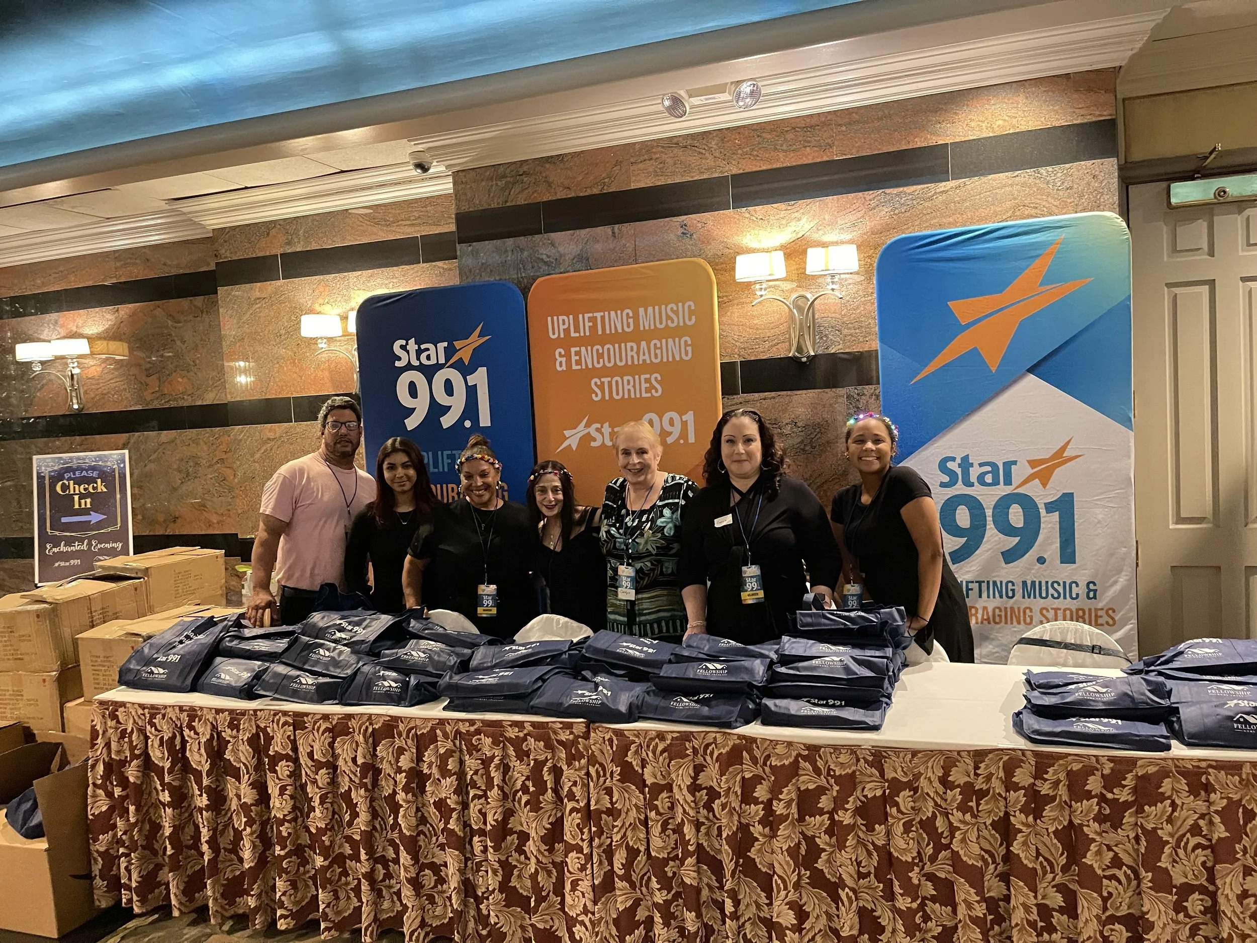 Group of six people standing behind a table filled with navy blue bags at a promotional event for Star 99.1 radio station, with a backdrop displaying the station's logo and branding.
