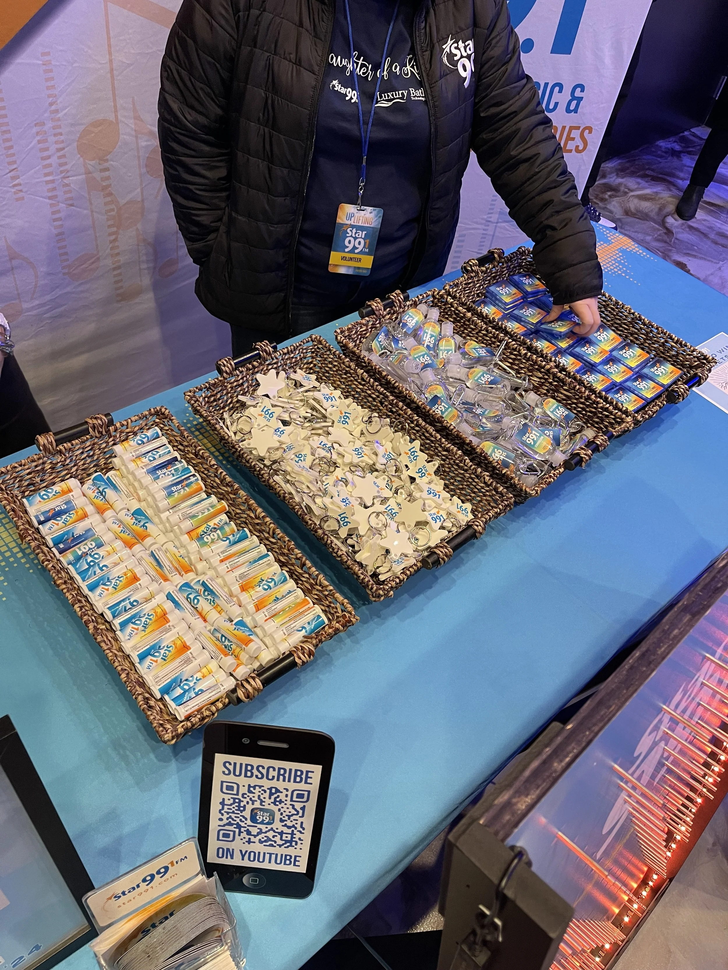 A person wearing a black jacket with a volunteer badge standing behind a table filled with promotional items, including lip balm, keychains, and small bottles, all branded with the Star 99.1 logo. The table is covered with a blue cloth, and there is 