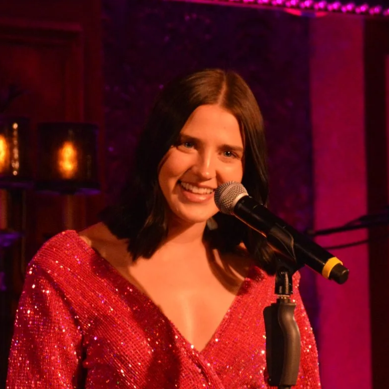 A woman with shoulder-length dark hair singing into a microphone on a stage with warm lighting.