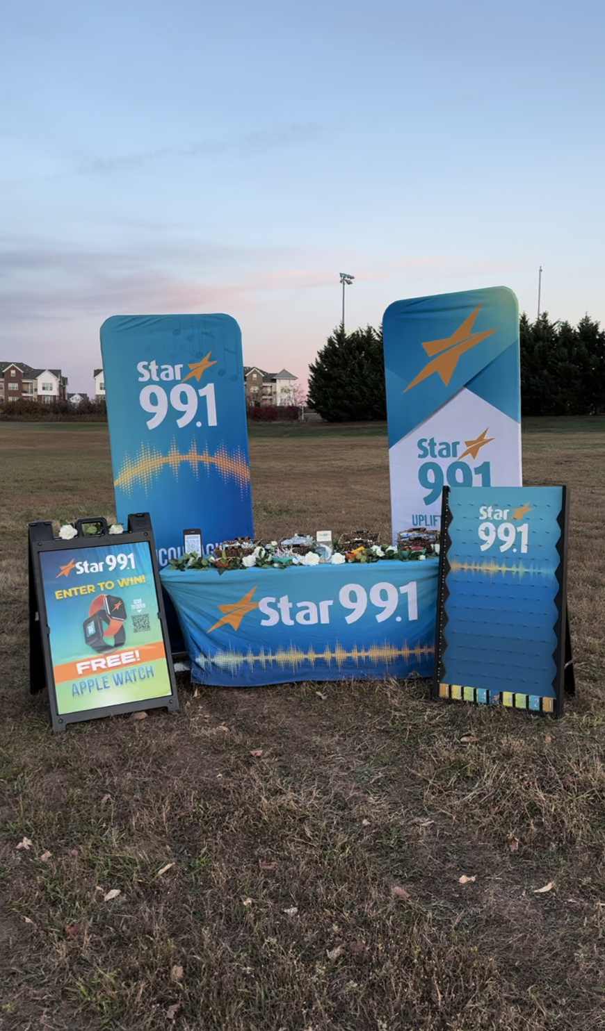 Outdoor setup for Star 99.1 radio station with promotional banners, flower decorations, and a sign offering a chance to win a free Apple Watch near houses and trees.