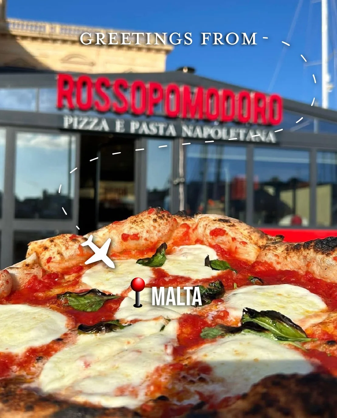 Close-up of a margherita pizza with melted cheese, basil leaves, and tomato sauce in front of a restaurant with a sign that reads 'Rossopomodoro Pizza e Pasta Napoletana' in Malta, with clear blue sky and a building in the background.