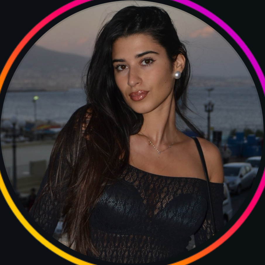A woman with long dark hair in a black bikini top with sequins, standing outdoors by the water during sunset, with hills in the background.
