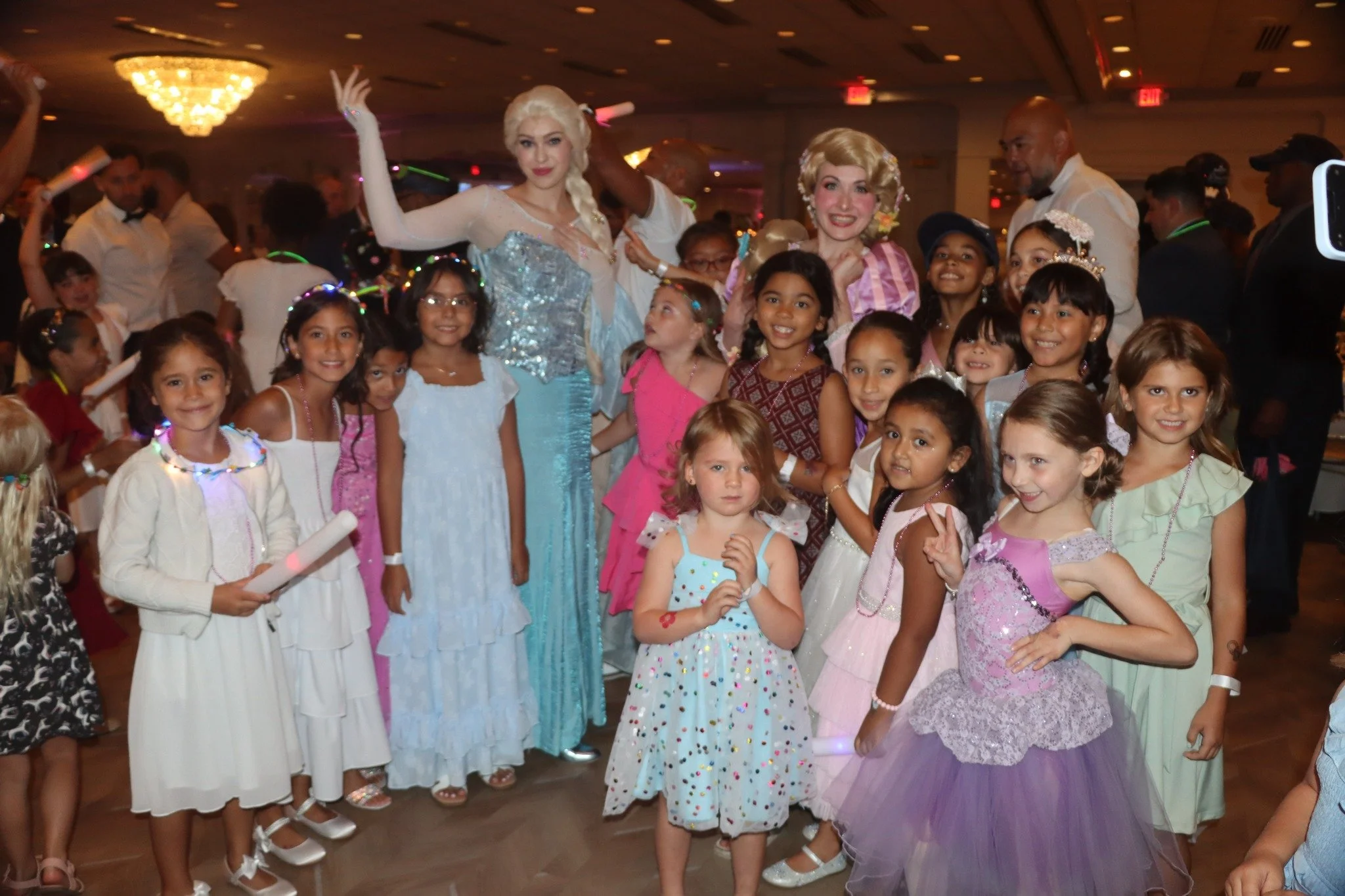 Children and adults dressed in costumes and fancy dresses at a party, with two women in princess costumes in the center.