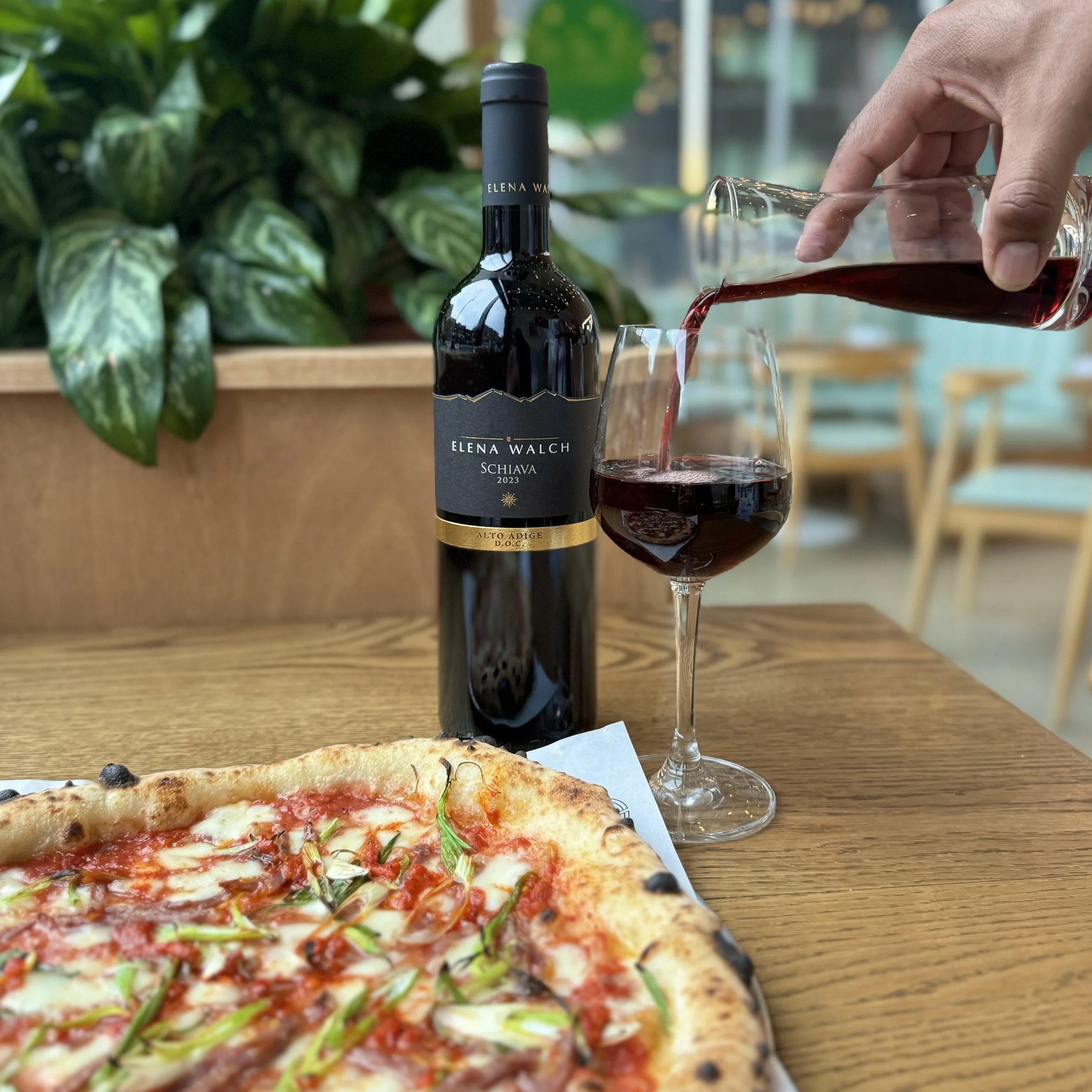 A bottle of Elena Walch Schiava 2023 red wine, a glass being filled with red wine, a pizza with cheese, tomato sauce, and green herbs on a wooden table, with leafy green plants in the background.