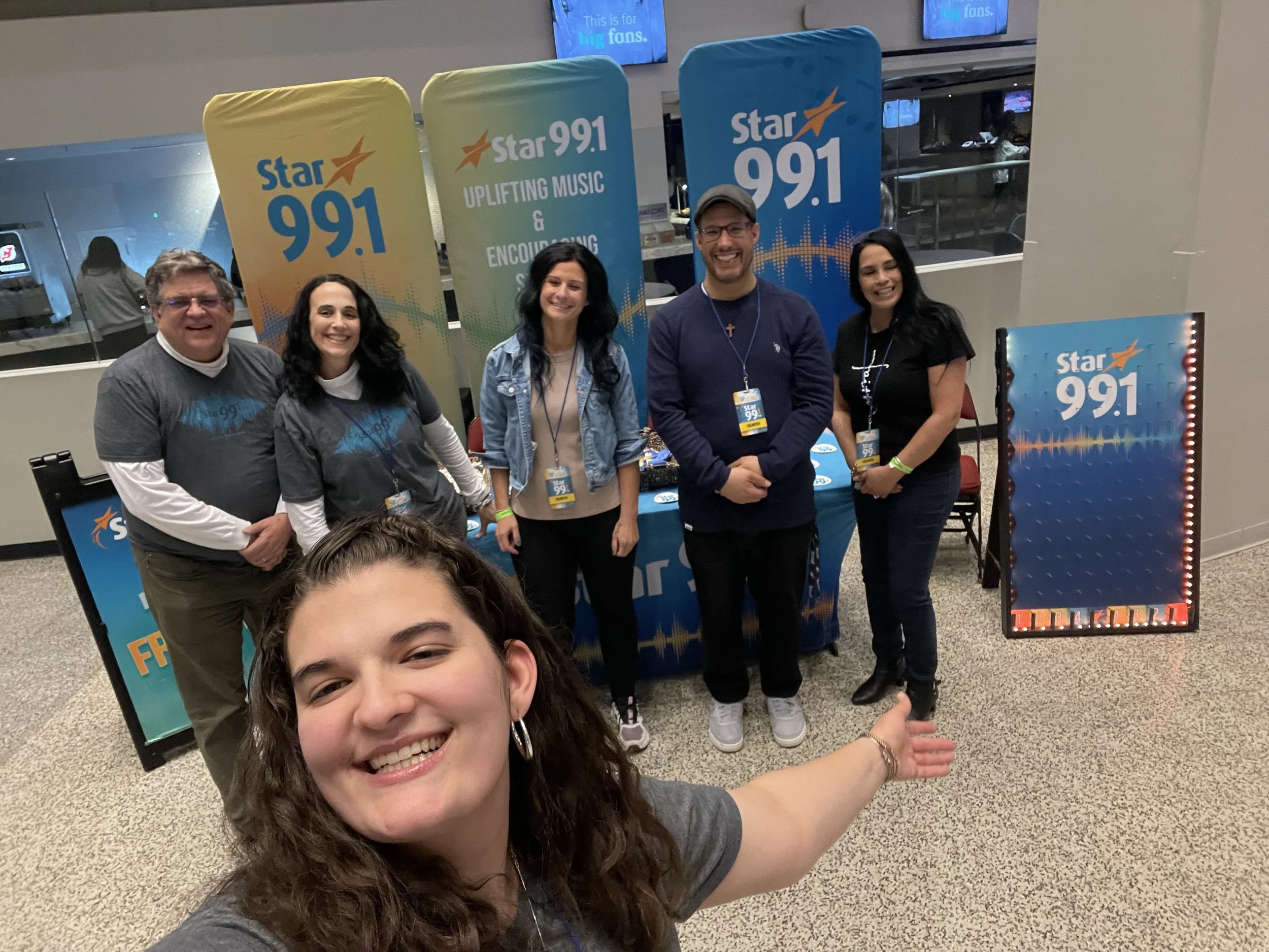 A group of six smiling people, five in the background and one in the foreground, posing in front of a billboard display for Star 99.1 radio station. The people are wearing casual clothes and some have station badges. The background includes several S