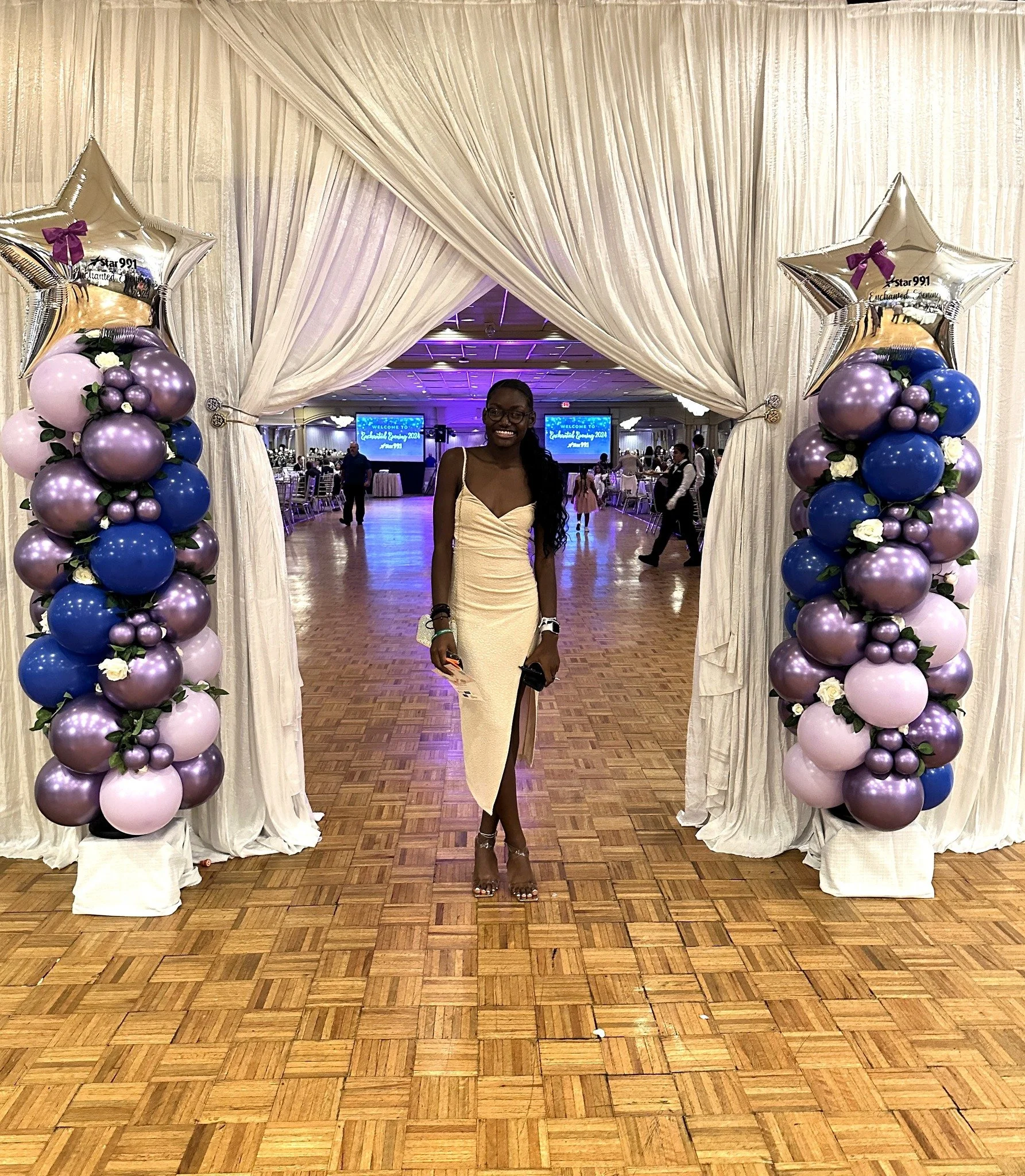 A woman in a beige dress standing in front of a decorated arch at a formal event or celebration.