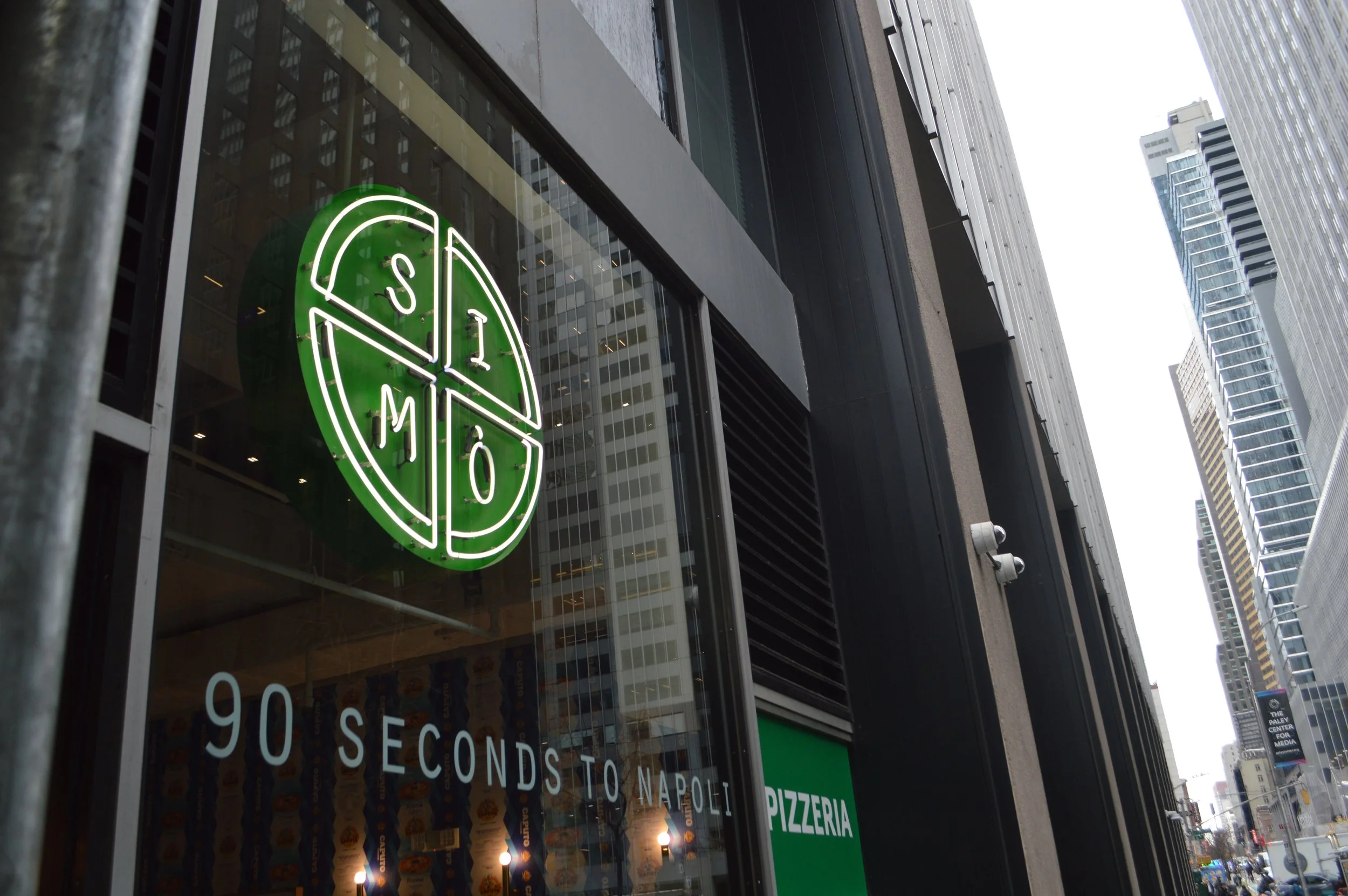 A neon sign of a pizza slice with the word 'SIMÒ' inside it, hung outside a restaurant on a city street. In Midtown, Manhattan. 