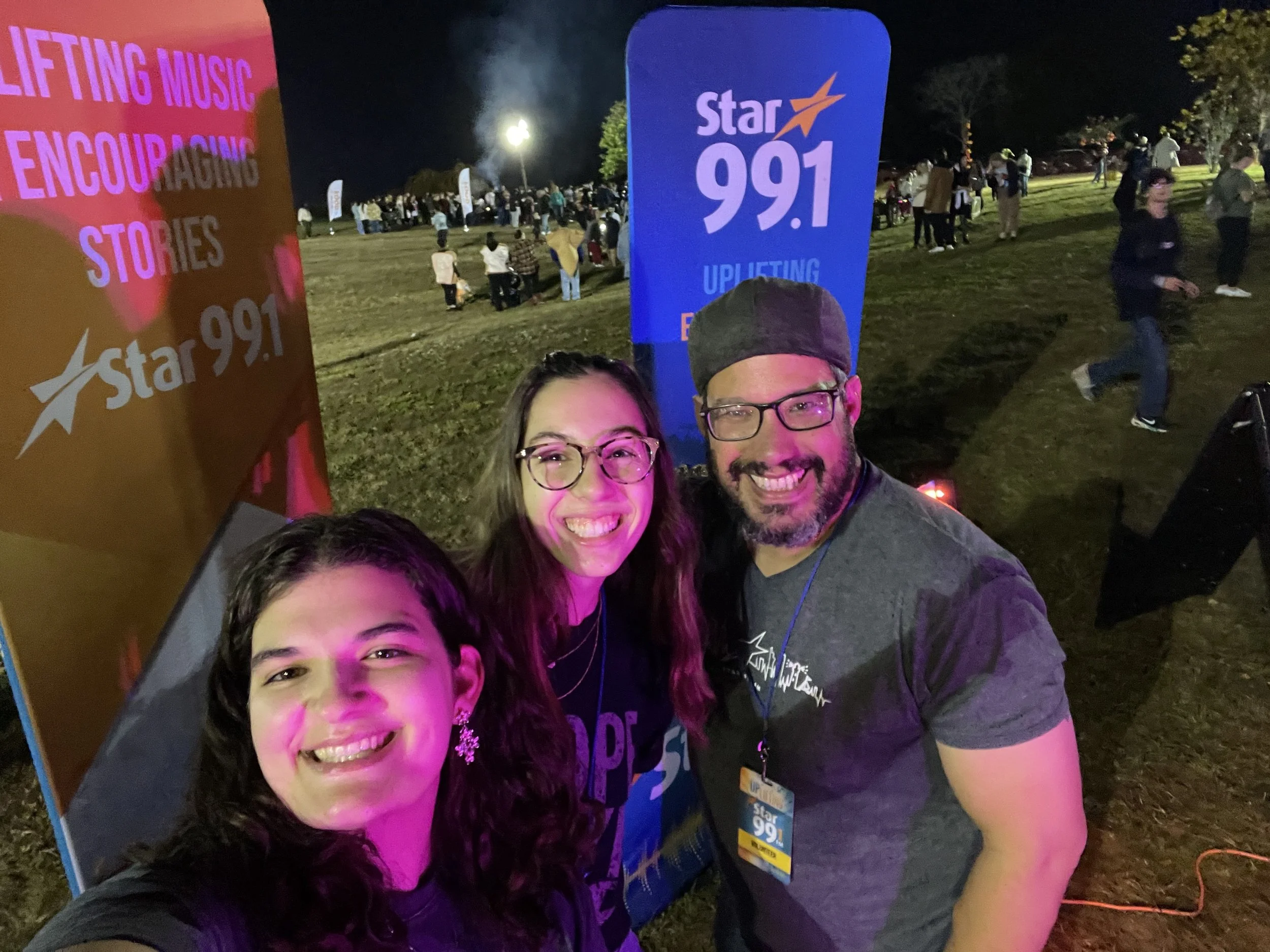 Three people smiling and taking a selfie at a nighttime outdoor event with a 'Star 99.1' radio station banner in the background, surrounded by other attendees and event signage.