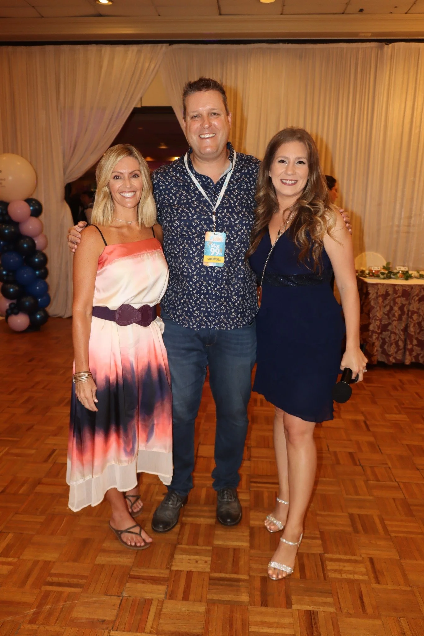 Three people standing close together at an indoor event, smiling at the camera. The woman on the left wears a colorful, sleeveless dress with a large belt and sandals. The man in the middle wears a patterned shirt with a conference badge around his n