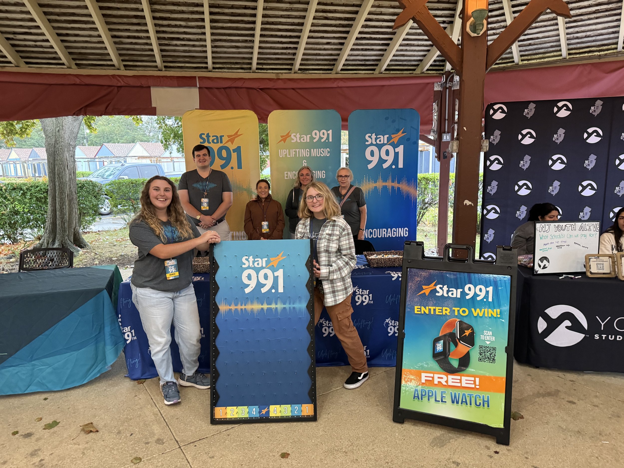 Group of five people standing behind a table at a promotional event for Star 99.1 FM radio station. Two women are in front holding a large blue sign with the station’s logo, while three are behind the table with promotional banners. The scene is outd