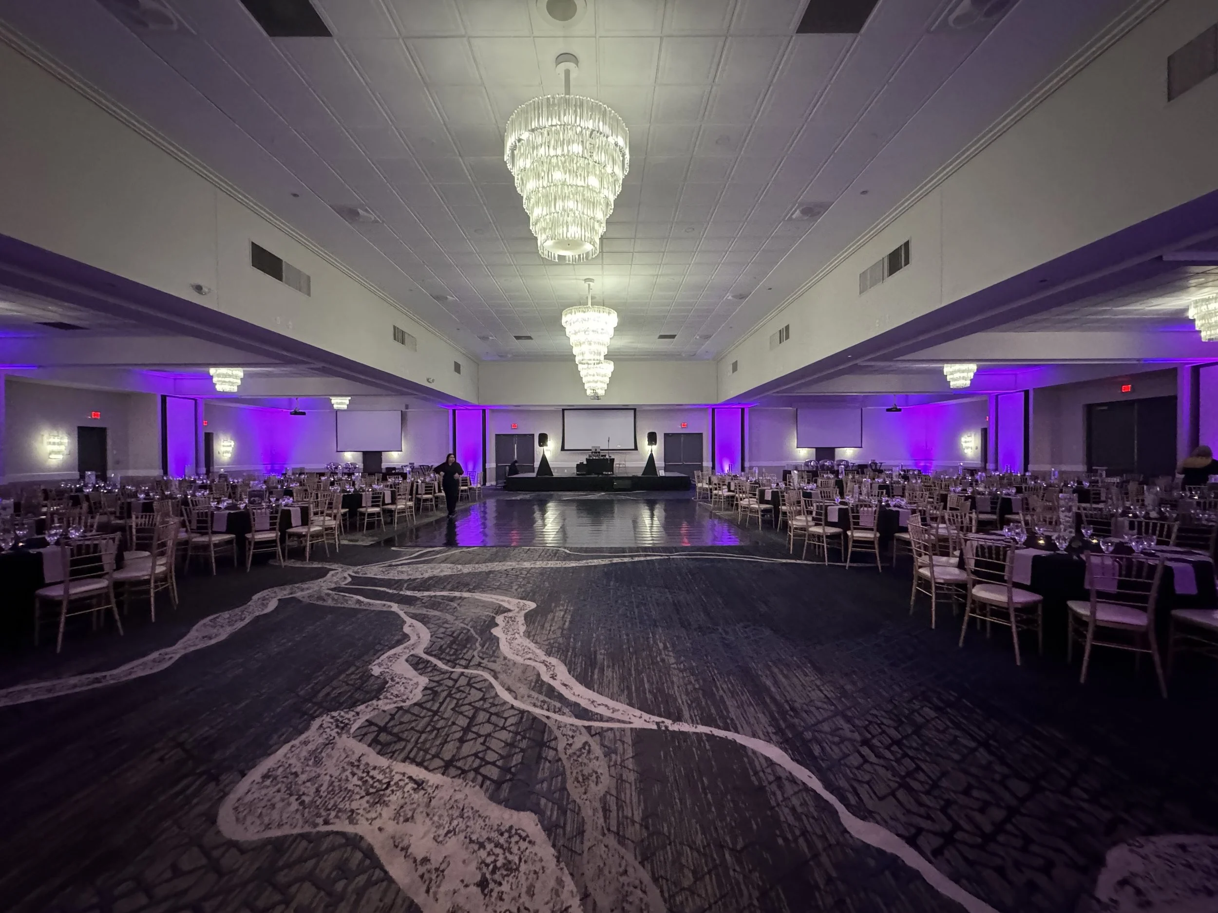 Empty banquet hall with round tables and chairs, a large chandelier hanging from the ceiling, purple lighting along the walls, a dance floor in the center, and a stage at the back with a screen and sound equipment.