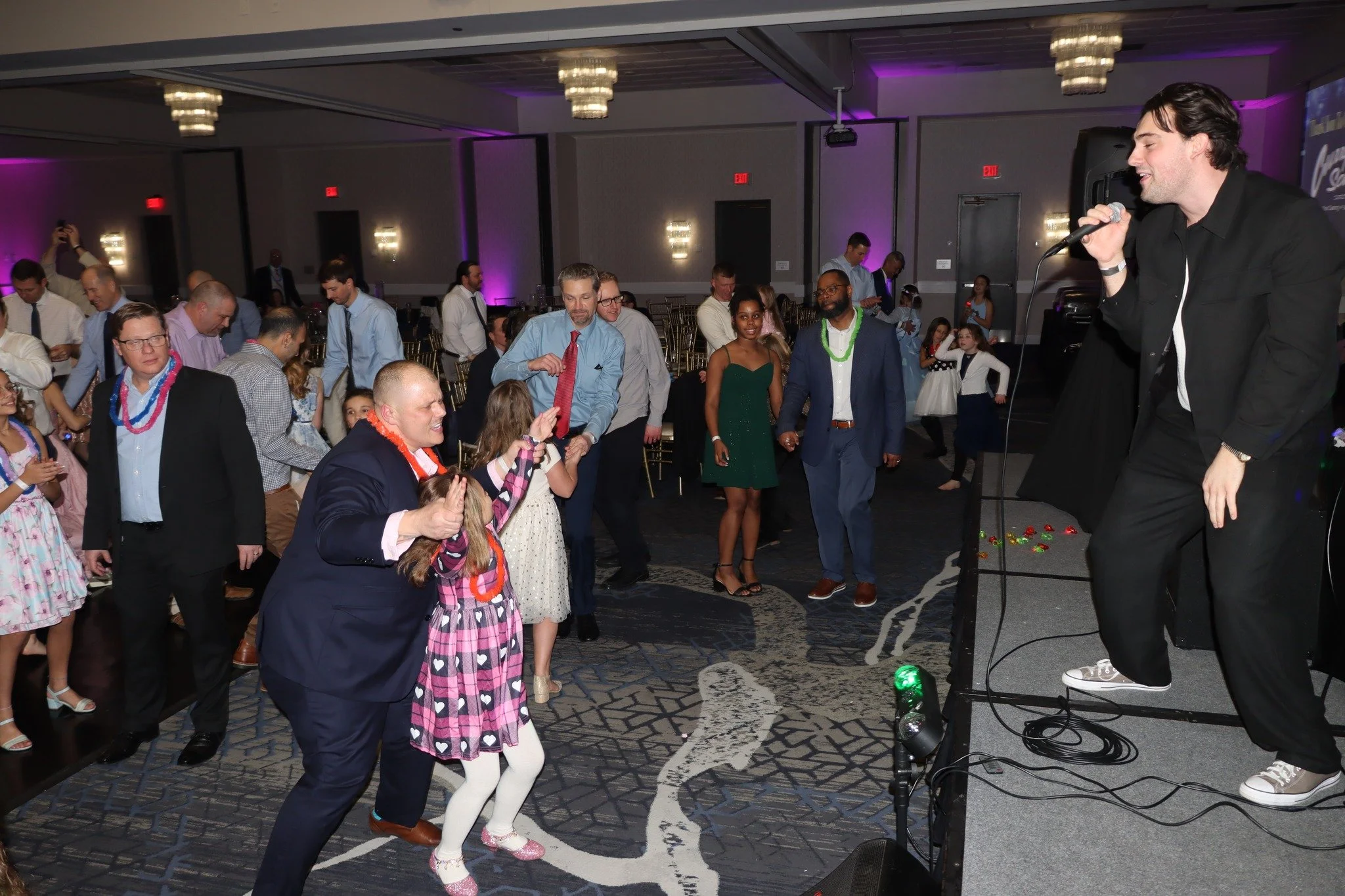People dancing and enjoying music at a party or celebration in a large, dimly lit event hall with purple lighting.