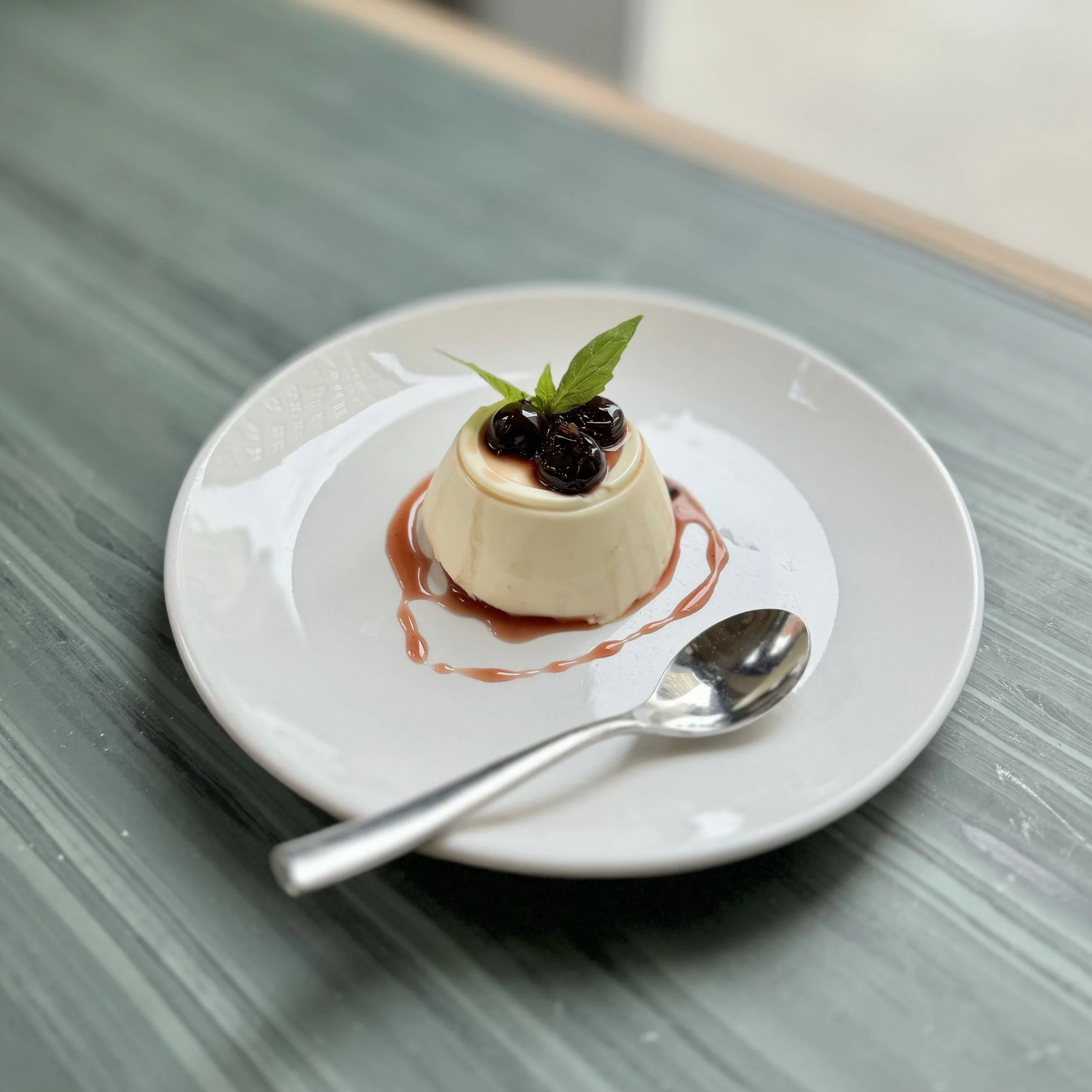 Creamy panna cotta topped with berries and a mint leaf on a white plate, with a silver spoon resting on the side, placed on a gray-green textured surface.