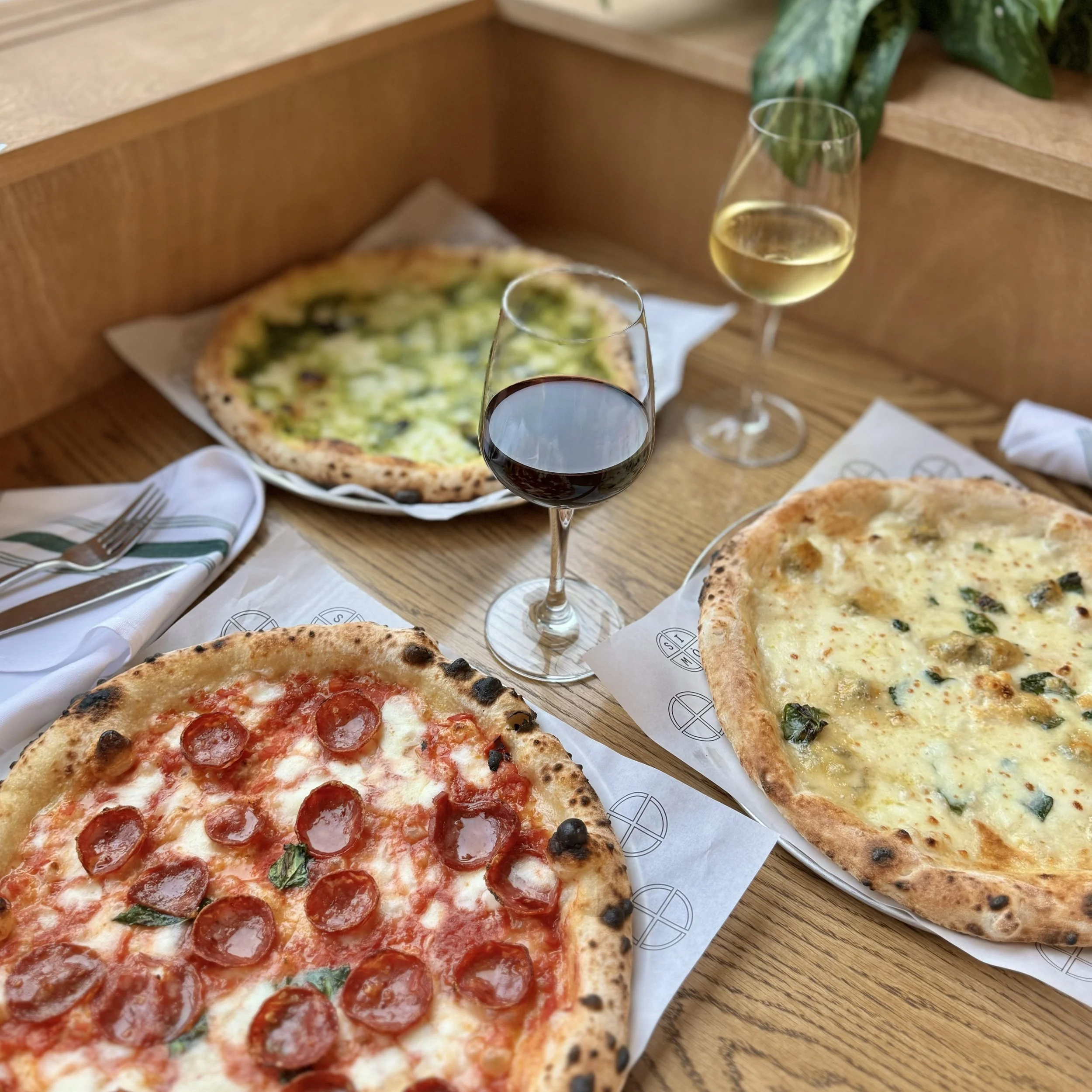 Three pizzas on wooden table with two glasses of wine, one red and one white, in a restaurant setting.
