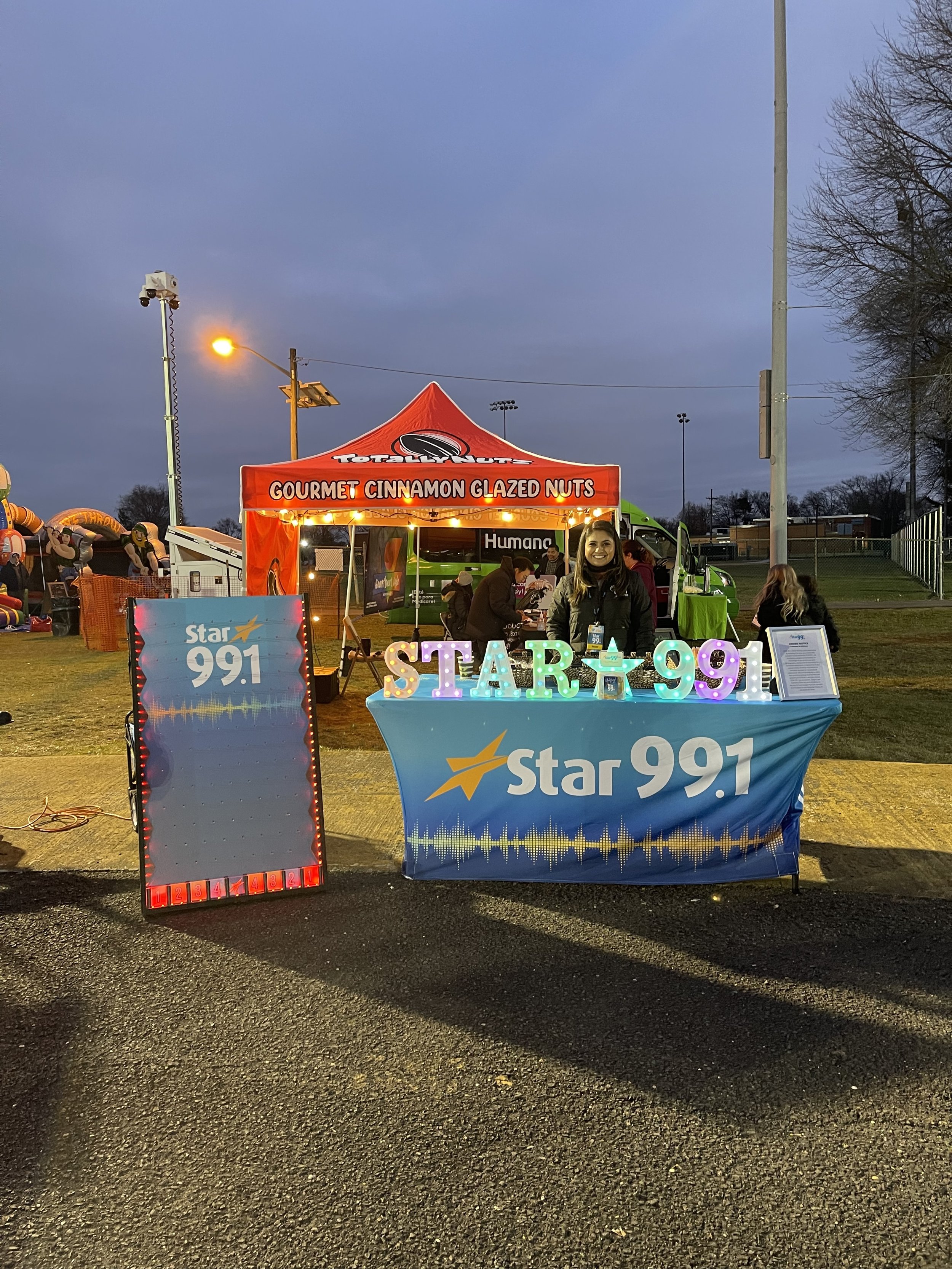 A booth for Star 99.1 radio station at an outdoor event, decorated with colorful lights and a sign displaying the station's logo, with a red tent behind and a woman smiling in front. There is a large gray and blue board with the station's logo to the